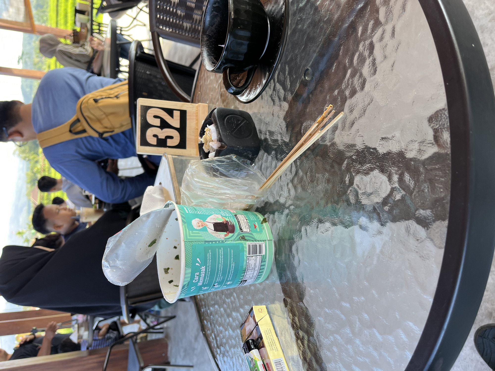 The image depicts a glass-top table at an outdoor seating area, possibly a restaurant or cafe, with a view of lush green rice fields in the background. The table is cluttered with various items. In the foreground, there is a half-open cup of instant noodles, a pack of cigarettes, a black ashtray with cigarette butts and plastic wrap, a black coffee cup and saucer, and a pair of chopsticks resting on the table. A sign with the number "32" is visible behind the noodle cup. Several people are partially visible in the background, including a person wearing a black hijab and another in a blue shirt with a yellow backpack. The overall impression is that of a casual meal or break being taken outdoors. The location is identified as Oro-oro Ombo, Indonesia, suggesting a tropical or sub-tropical setting.