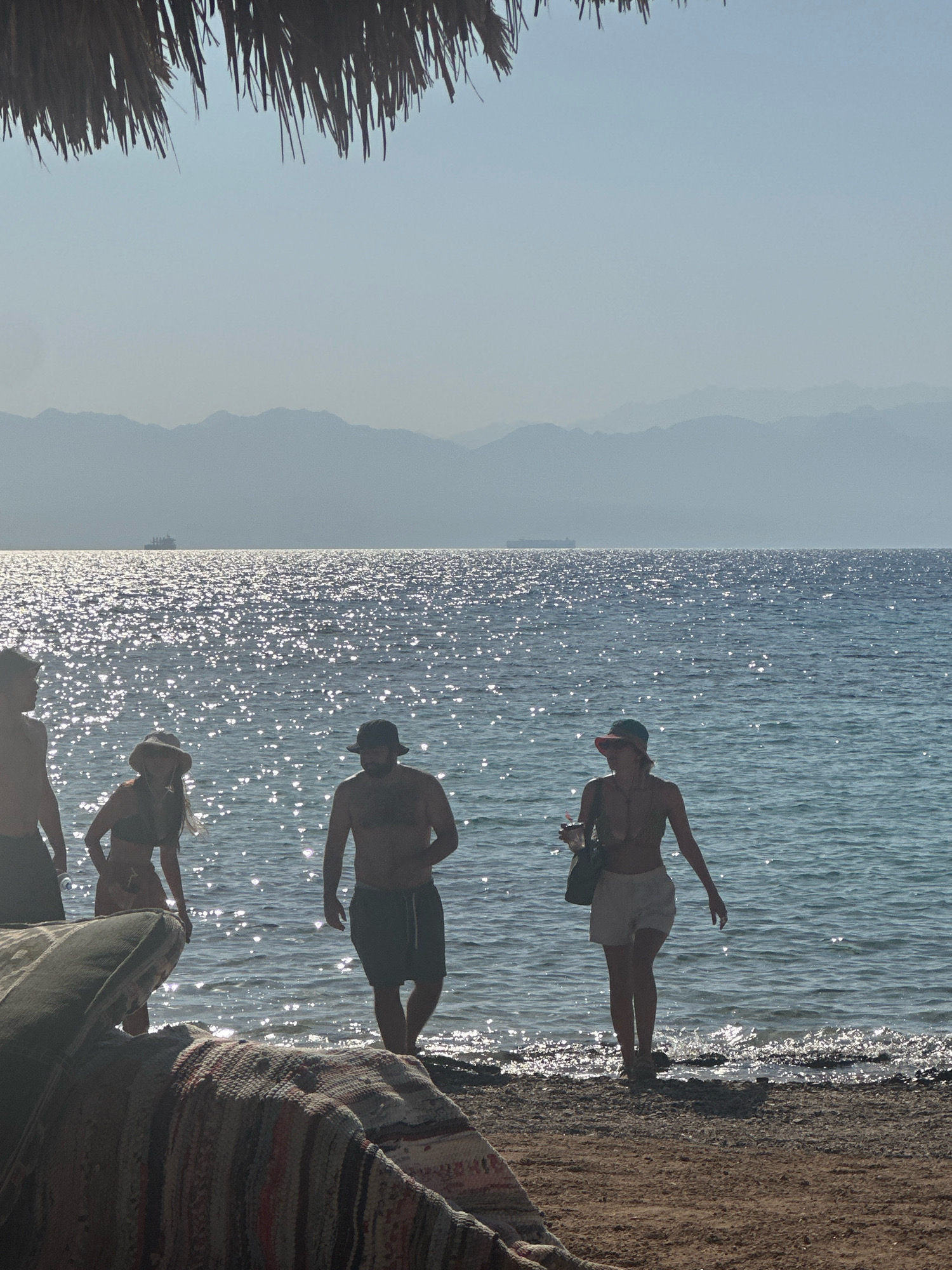The image depicts a scene at a beach in Nueiba, Egypt, under a clear sky. In the foreground, the edge of a woven rug or blanket is visible, partially obscuring the sandy shore. The water of the sea is bright with sunlight reflecting off its surface, creating a shimmering effect.
Several people are present near the water's edge. On the left, a person, silhouetted and only partially visible, stands facing the sea. To their right, a woman wearing a wide-brimmed hat and a bikini is walking towards the shore. She appears to be holding a drink in her right hand. Further to the right, a man wearing a hat and swim trunks is walking towards the sea. Another person, a woman in a bikini and shorts, is walking away from the sea, carrying a bag and a drink.
In the middle ground, a ship is visible on the horizon, and in the background, a range of hazy mountains stretches across the landscape, characteristic of the Sinai Peninsula in Egypt. The overall atmosphere is serene and suggests a leisurely day at the beach.
