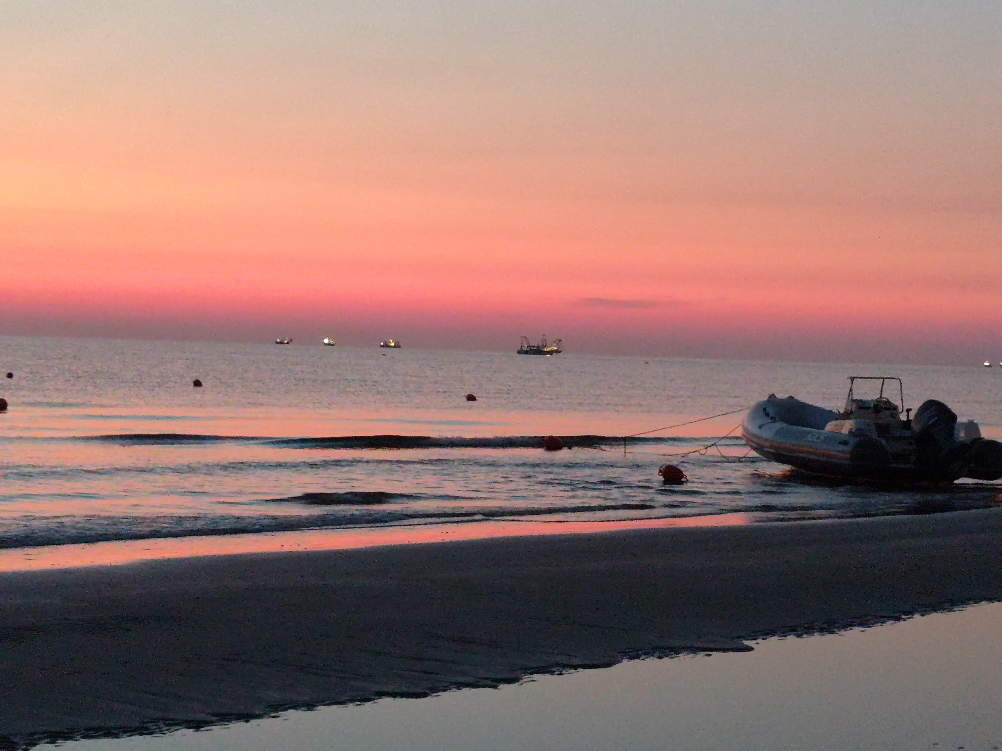 A wide-angle, low-angle shot captures a serene beach scene at sunset in Rimini, Italy. The sky transitions from a pale yellow at the horizon to a vibrant pink and then a soft lavender as it ascends. Several small fishing boats with illuminated lights dot the calm sea in the distance. In the foreground, a rigid inflatable boat (RIB) with twin outboard motors rests partially on the sandy shore, tethered to a red buoy. Another red buoy floats nearby in the gentle waves. The water reflects the warm hues of the sunset. The beach itself is dark and wet, with the tide receding. The overall impression is one of tranquility and the end of a day at the coast.