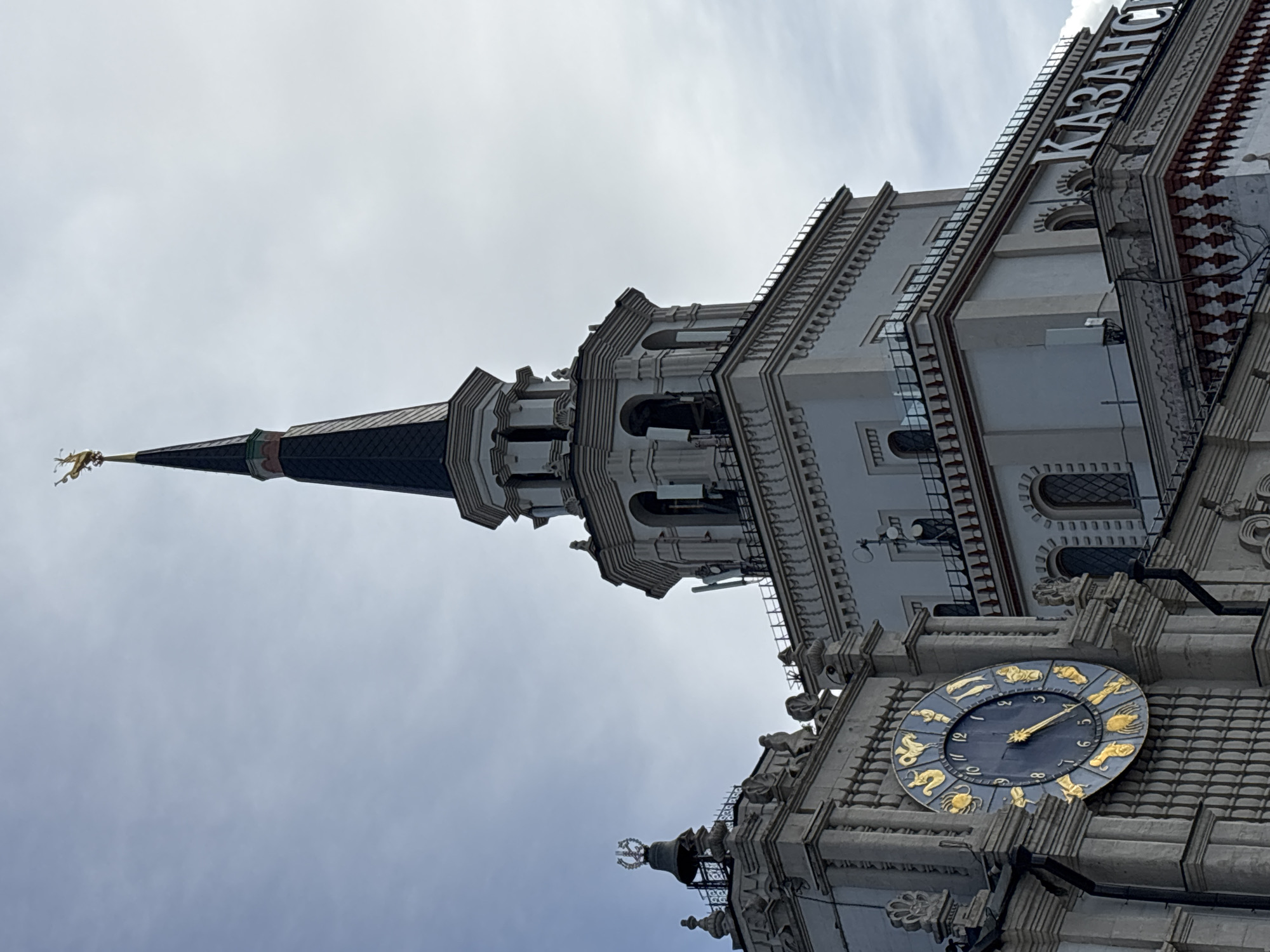 The image shows the clock tower of the Kazan railway station in Moscow, Russia. The tower features a large zodiac clock, a spire topped with a golden figure, and the word "KAZAN" written vertically on the building. The sky is overcast. The image is taken from a low angle.