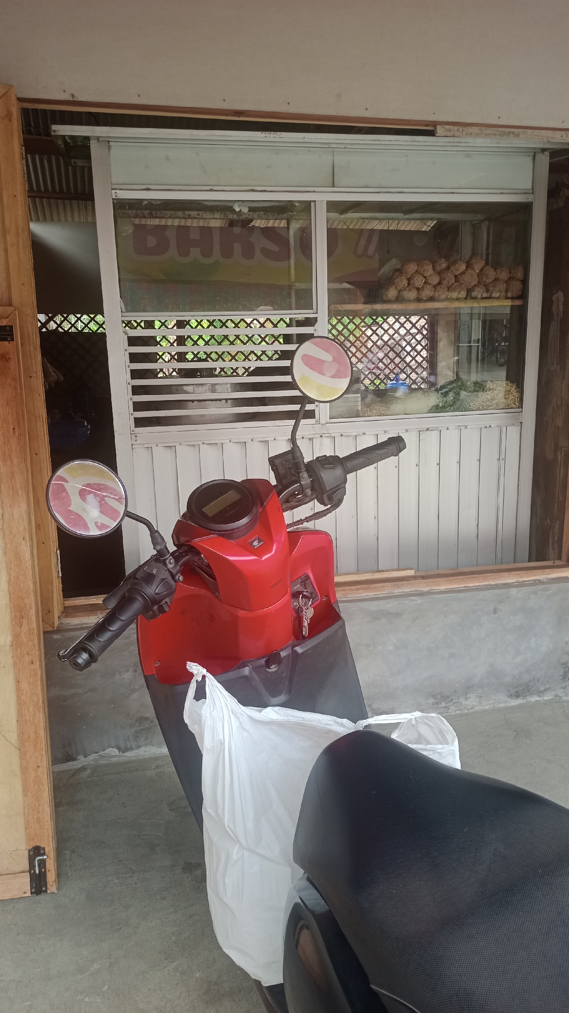 A red scooter is parked in front of a small storefront in Cikalong, Indonesia. The storefront has a large window displaying food items, including what appears to be meatballs and vegetables. A white plastic bag is hanging from the scooter's handlebars. The ground is concrete.