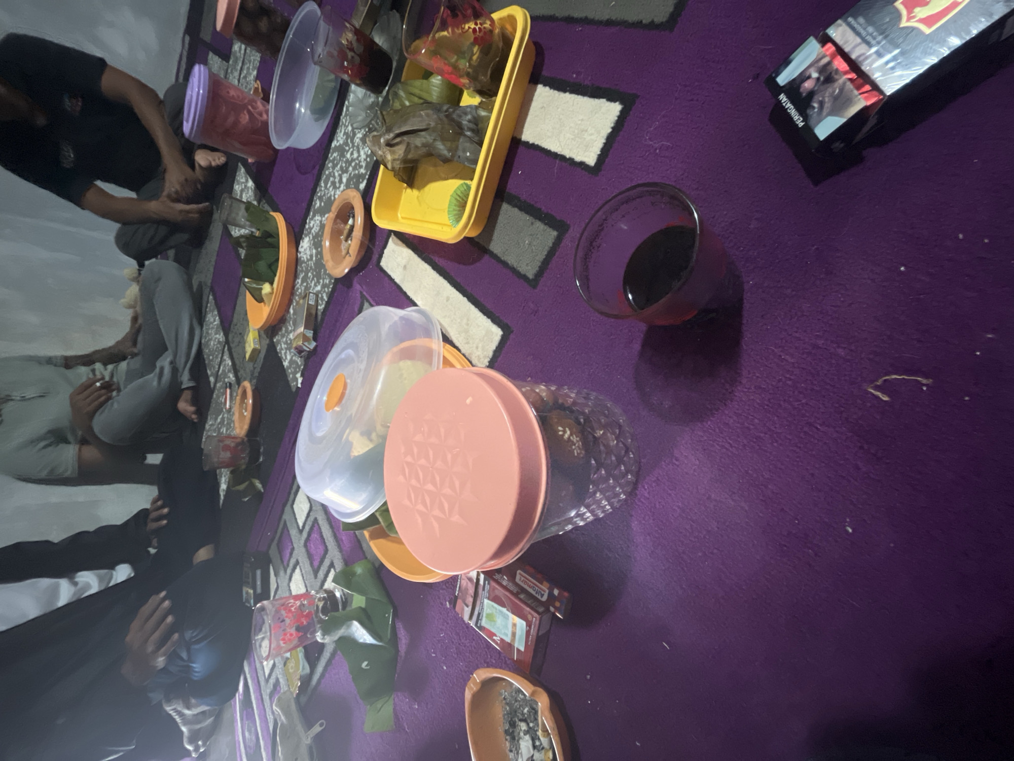 In Karang Sari, Indonesia, a group of individuals is seated on a purple carpeted floor, likely a living room or gathering space, around an assortment of food and drinks. The scene depicts a casual, informal gathering, possibly a meal or a break.
Several individuals are visible, some partially obscured. One person on the left wears a dark outer garment, while another in the center wears a light gray t-shirt and pants. A third person on the right is in a dark t-shirt. Their postures suggest they are relaxed and engaged in conversation or simply resting.
The floor is covered with a purple carpet with a geometric pattern of beige and dark gray squares. Scattered across the carpet are various objects related to consumption. There are several glasses, some containing a dark liquid, and others appear empty or with remnants of drinks. A large, clear plastic container with a pink lid sits prominently in the foreground, containing what appears to be food. Another container with a purple lid and red contents is visible further back.
Food items are present in a yellow rectangular tray, including what looks like wrapped parcels of food. There are also several packets of cigarettes, one of which is open and showing its contents. An ashtray filled with cigarette butts is also on the floor. A plastic lid, likely from a food container, is also present. A closed cigarette pack with "PERINGATAN" (Indonesian for "WARNING") printed on it is visible in the bottom right corner of the image.
The lighting in the room appears dim, casting shadows and creating a somewhat subdued atmosphere. The overall impression is of a relaxed social gathering where food, drinks, and smoking are part of the experience.