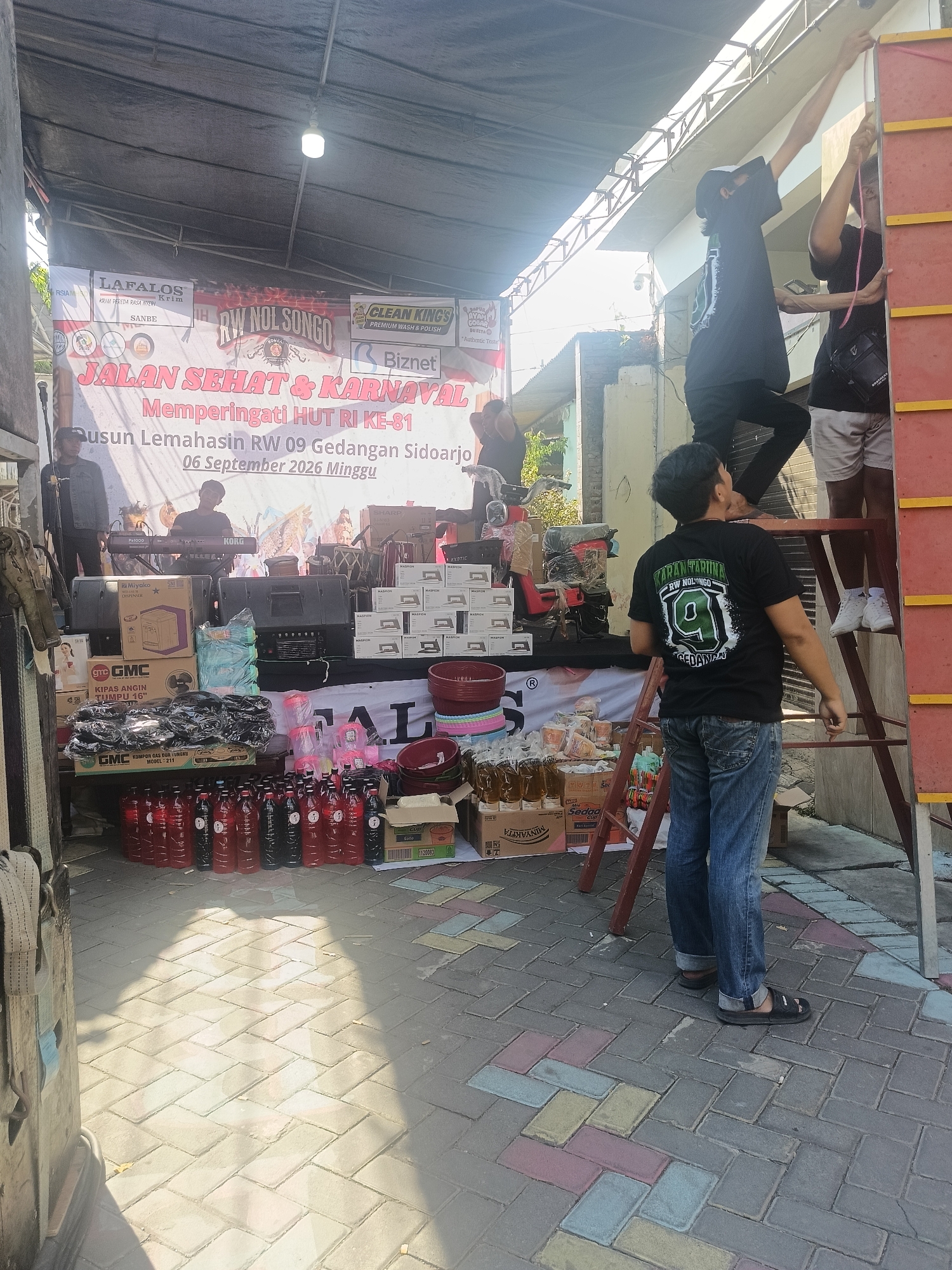 The image depicts an outdoor scene, likely in Gedangan, Indonesia, featuring a temporary stage setup for an event. A banner is prominently displayed, reading "JALAN SEHAT & KARNAVAL Memperingati HUT RI KE-81 Dusun Lemahasin RW 09 Gedangan Sidoarjo 06 September 2026 Minggu." This translates to "Healthy Walk & Carnival Commemorating the 81st Anniversary of the Republic of Indonesia, Lemahasin Hamlet RW 09 Gedangan Sidoarjo, September 06, 2026 Sunday."
Several individuals are present. Two men are seen on a ladder, one appears to be hanging a rope. Another man is standing on the ground, looking up at them. In the background, two men are positioned near what seems to be a keyboard and sound equipment, suggesting a musical performance.
The scene is filled with various items, including boxes of merchandise, plastic bottles filled with red liquid, stacks of red and blue plastic bowls, and an assortment of electronic equipment like speakers and a fan. The ground is paved with interlocking bricks in varying shades of gray, red, and yellow, with dappled sunlight casting shadows. The overall impression is of a community event being prepared or underway.