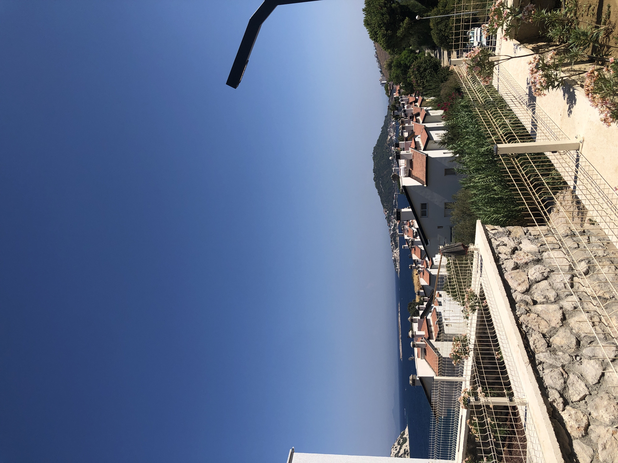 The image shows a scenic view of the city of Karaburun, Turkey, with the deep blue Aegean Sea in the background. The foreground features a stone wall topped with a white concrete ledge and a light-colored metal fence. Beyond the fence, there is a paved walkway lined with green vegetation and blooming pink flowers. A row of white houses with terracotta roofs stretches along the coastline, interspersed with trees and other foliage. In the distance, a prominent green hill rises from the landscape, dotted with buildings. A modern black streetlamp extends into the upper right corner of the frame. The sky is a clear, vibrant blue. A white vehicle is visible on a road in the middle ground.