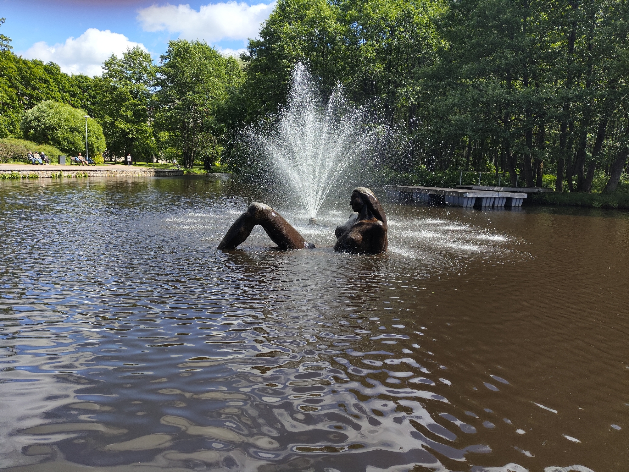 The image depicts a bronze sculpture of a reclining nude woman partially submerged in a lake in Tallinn, Estonia. The sculpture is positioned in the foreground, with a fountain spraying water in the background. The lake is surrounded by lush green trees and a park area with people sitting on benches. The sky is blue with scattered white clouds. The scene appears to be a public park on a sunny day.