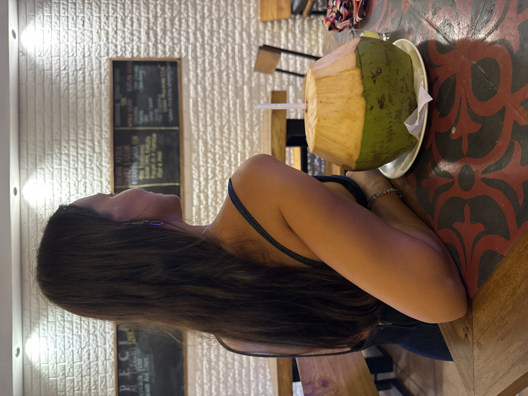 The image shows a woman seated at a patterned table in a casual dining setting, likely in Kuta Selatan, Indonesia. She is seen from a side profile, with long, dark brown hair flowing over her shoulder and arm. She wears a dark top with thin straps, and her left arm rests on the table, adorned with a bracelet. Her attention seems directed towards the left side of the frame.
In front of her, on the dark red and black patterned tabletop, is a fresh coconut drink served on a white plate with a napkin and a clear straw. The coconut is partially husked, showing a lighter, yellowish-green base and a darker green top.
The background features a textured white brick wall, illuminated by recessed ceiling lights, suggesting an indoor or evening setting. On the wall, a dark blackboard menu with a wooden frame is visible. Although most text is blurry due to the shallow depth of field, words such as "SPECIAL" and "MIXED GRILL" can be faintly made out, indicating a food and drink establishment. The overall scene conveys a relaxed atmosphere.