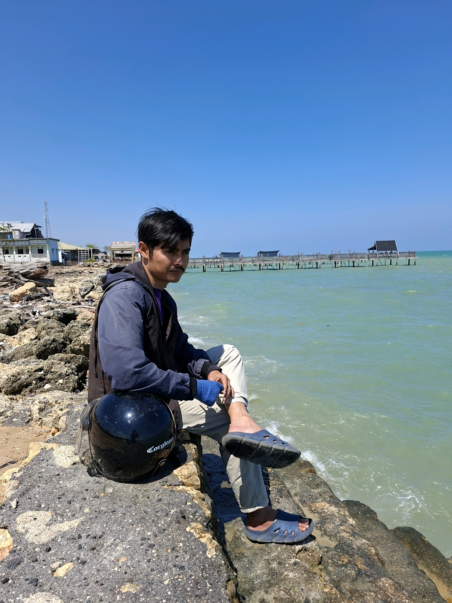 A young adult male sits on a rocky shoreline in Sejati, Indonesia. He is wearing a dark blue jacket over a purple shirt, tan pants, blue gloves, and dark blue sandals. A black motorcycle helmet rests on the rock in front of him. The background features a calm, light green ocean with a wooden pier extending into the water, and several buildings on the distant shore. The sky is clear and blue.