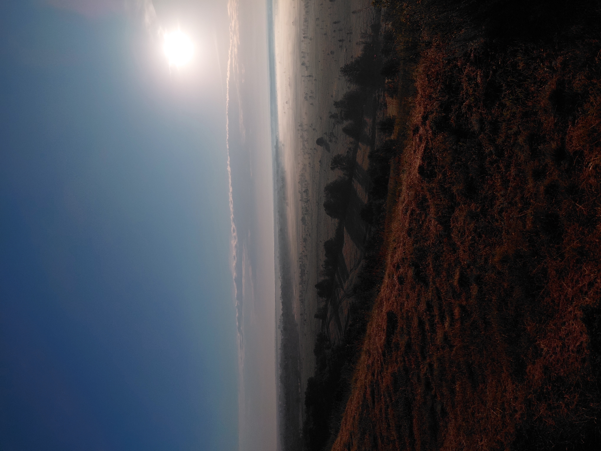 This is a wide-angle, low-angle shot of a misty, rural landscape at dawn, taken from an elevated position on a grassy hill. The foreground is dominated by the reddish-brown, dry grass of the hill. The middle ground reveals a valley with scattered trees and fields shrouded in a dense layer of fog, obscuring the ground details in many areas. The far distance shows a hazy horizon with what appears to be a small town or village. The sky is a gradient of clear, deep blue at the top, transitioning to lighter blues and then a pale, hazy white near the horizon where the sun is intensely bright, partially obscured by thin clouds. The overall atmosphere is serene and slightly melancholic, characteristic of early morning in a temperate climate. The location is specified as Ustyvytsia, Ukraine. There are no people or discernible actions in the image.