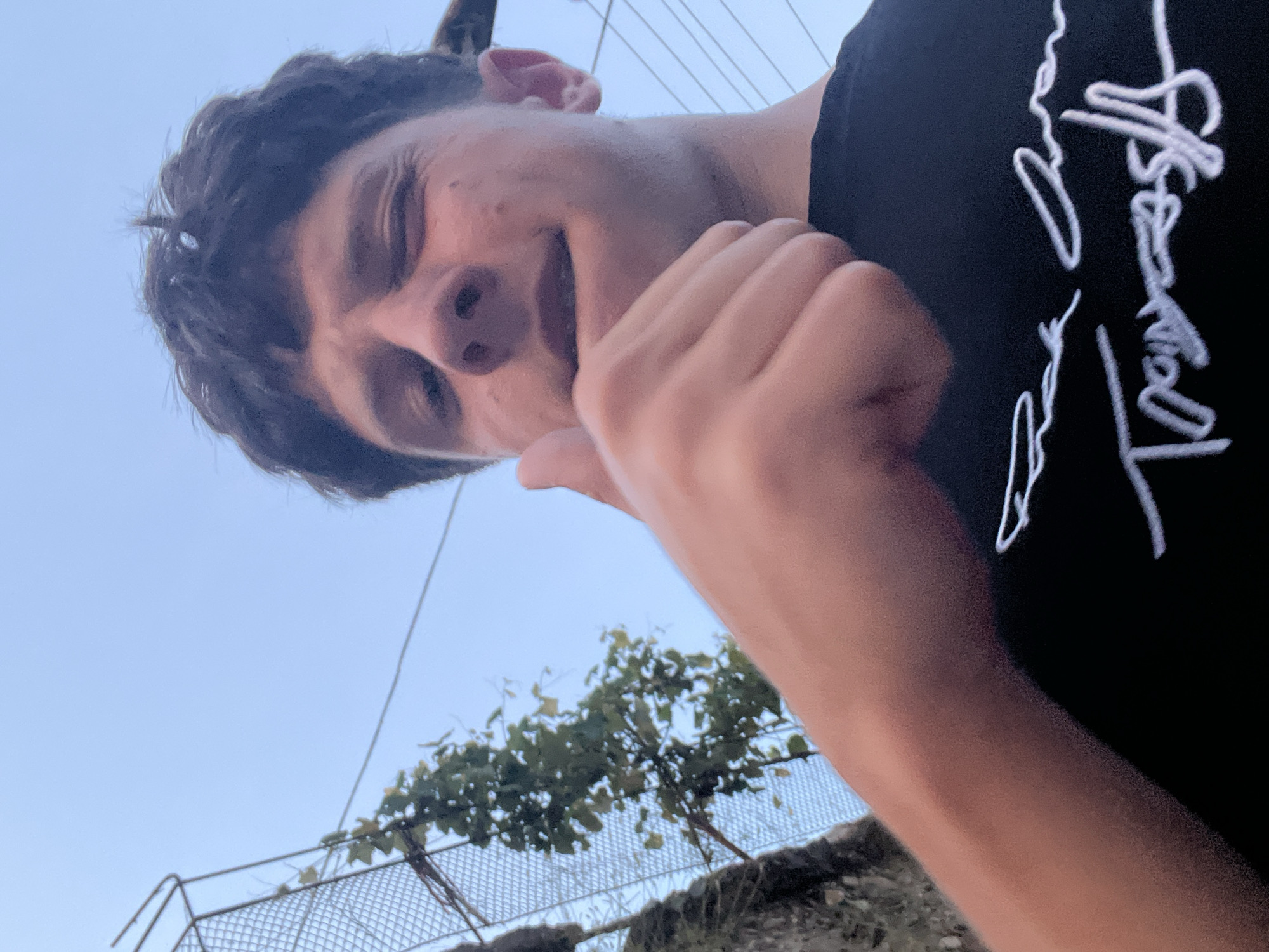 A young male subject with dark, disheveled hair and fair skin is depicted from a low-angle perspective, facing the camera. His right eye (viewer's left) is partially closed in a squint or wink, and his mouth is slightly open. He is wearing a dark-colored t-shirt with white, stylized, handwritten text or graphics across the chest, which partially reads "Traces" and other cursive elements. His right hand is raised, performing a "thumbs up" gesture, occupying the lower-center of the frame.
The background features a light blue sky, indicating an outdoor setting in Zlatograd, Bulgaria. To the left, a section of a metal mesh fence with a grid pattern is visible, alongside dark green foliage and tree branches with leaves. Faint power lines or cables are discernible against the sky in the upper right quadrant of the image. The overall lighting is soft and diffuse.