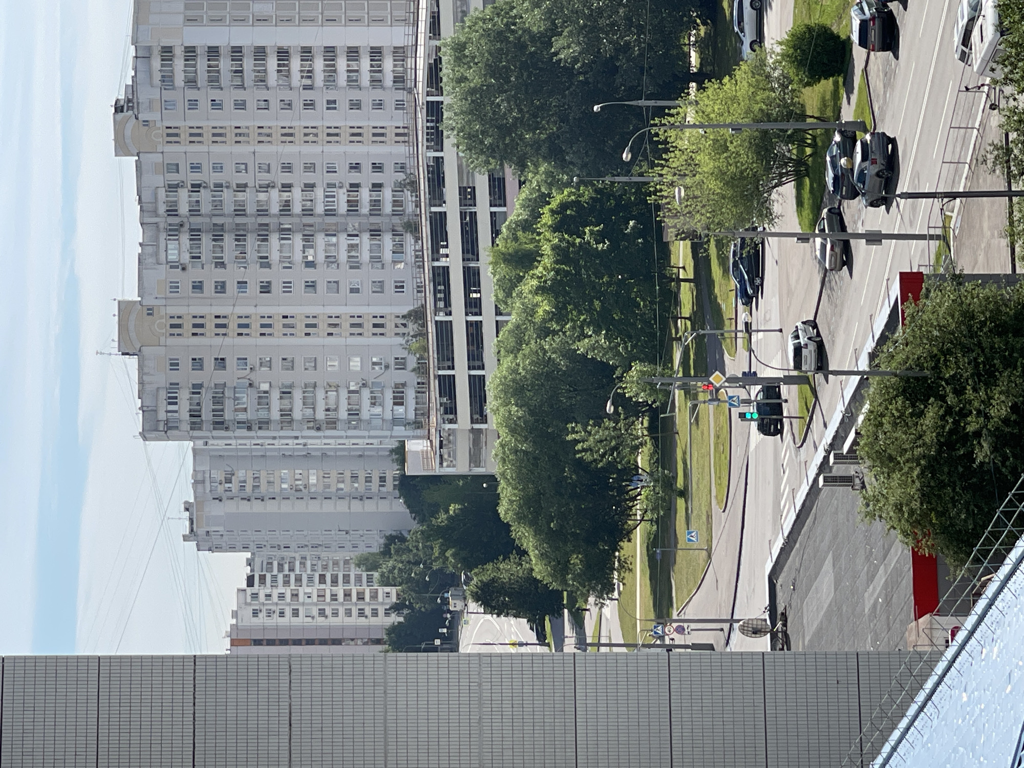 The image depicts a view of a residential area in Moscow, Russia, on a clear day. In the foreground, a portion of a modern building with a grey tiled facade is visible. Further back, a multi-story parking garage with a light-colored exterior stands prominently. Beyond that, a series of tall, light-colored apartment buildings with numerous windows line the horizon. Lush green trees are scattered throughout the scene, with a road network and several cars visible on the street. Traffic lights are also present, indicating an intersection. The sky is a pale blue with some wispy clouds.