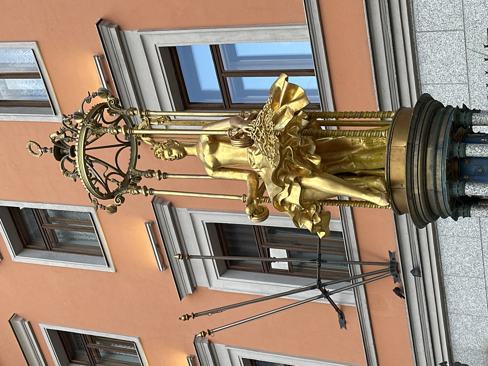 A golden statue of a woman holding an object, adorned with ornate golden cloth, is situated within a cylindrical, decorative metal enclosure. The enclosure is attached to a building with a salmon-colored facade and several framed windows. Attached to the building are metal rods or supports. The location appears to be Moscow, Russia.