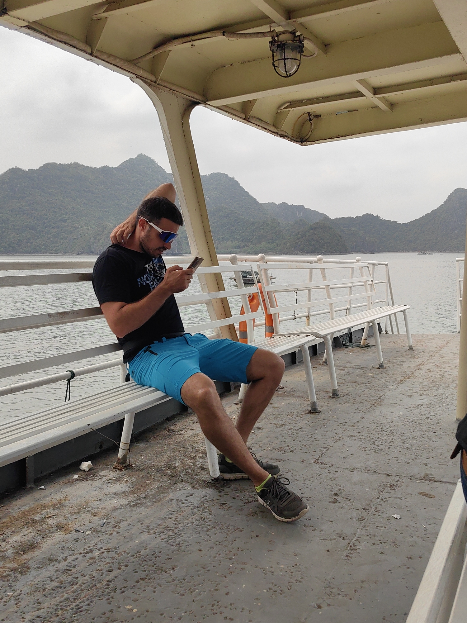 A man is sitting on a bench on what appears to be a ferry or boat. He is wearing a black T-shirt, bright blue shorts, and athletic shoes. He is looking at his mobile phone. He is wearing white-framed sunglasses. The boat has a metal roof and white benches with backs. In the background, there are mountains and water. The floor of the boat is metal and shows signs of wear and tear. A life preserver is strapped to the railing of the boat. The photograph was taken in Ha Long Bay, which is part of the city of Hải Phòng, Vietnam.