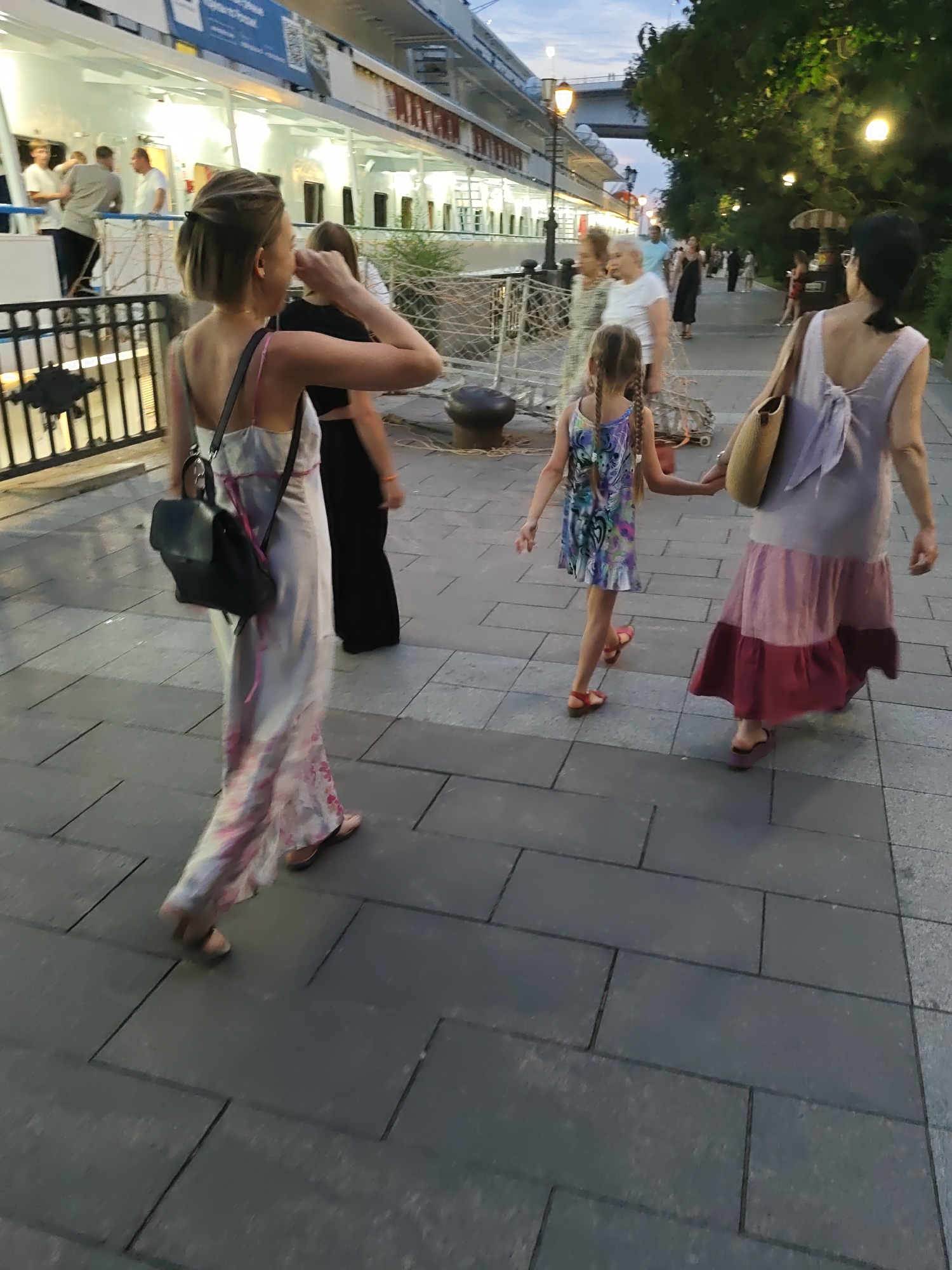 The image depicts a waterfront promenade in Rostov-on-Don, Russia, with a large white cruise ship docked to the left. The time appears to be late afternoon or early evening. In the foreground, a woman in a long, light-colored dress with floral patterns walks away from the camera, carrying a black backpack. To her right and slightly ahead, a young girl with braided hair in a colorful dress walks hand-in-hand with another woman. This second woman, further to the right and also walking away from the camera, wears a long, two-toned dress and carries a straw bag over her shoulder. Behind them, other individuals can be seen walking along the promenade. The promenade is paved with large, rectangular gray tiles. In the background, trees and streetlights line the walkway, and a bridge is visible in the distance. The scene conveys a sense of leisurely activity on a summer evening.