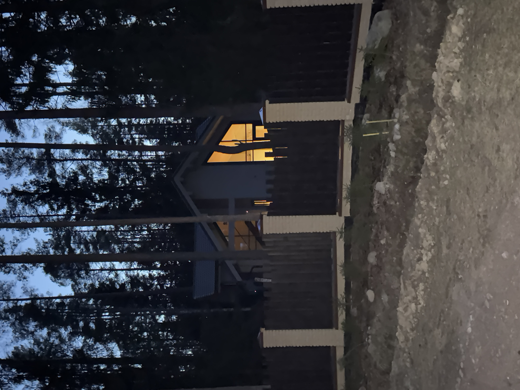 The image depicts a nighttime scene in what appears to be a wooded area, with a house visible through the trees and a fence. The house has a steeply pitched roof, and a window is brightly illuminated from within. The fence is made of wood with decorative post caps, bordering a dirt path or road. Tall, dark trees are silhouetted against a dusky sky. The image seems rotated, but the information about the location cannot be confirmed.