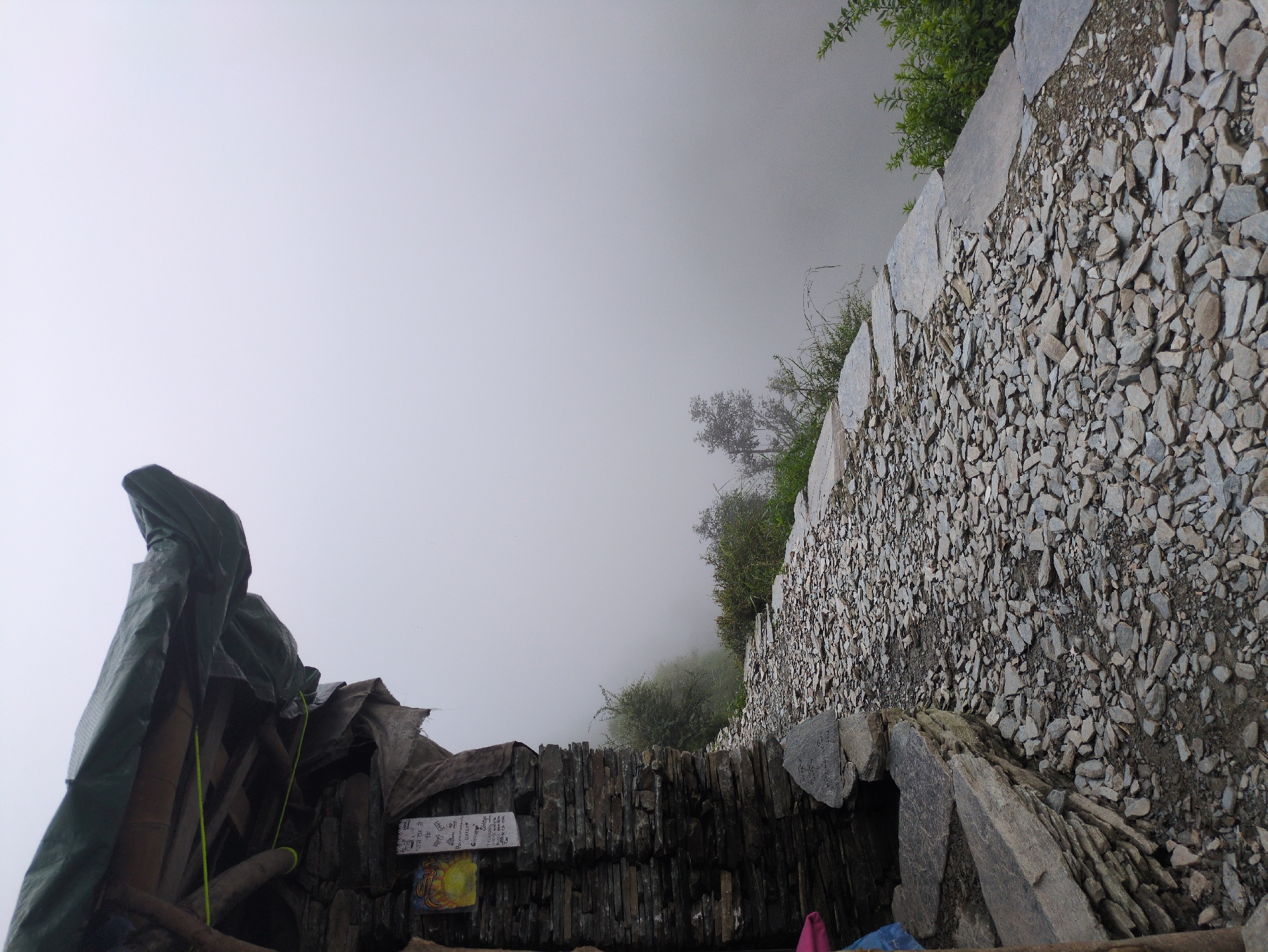 An outdoor scene shows a rocky path leading past a weathered stone structure with a tarp roof. The path is made of small, irregular grey stones, and some larger flat stones form a border along the right side. To the left, a section of a stone building is visible, constructed from stacked dark grey and brown flat stones. The building has a rough wooden frame supporting a dark green tarp that extends outwards to form an overhang. A yellow and red religious or informational sign is attached to the wall of the building, with some text below it. Green vegetation is visible along the edge of the path and further in the distance. The background is obscured by thick white fog, giving the impression of a high-altitude or misty environment. The scene appears to be a pathway in a mountainous region, possibly a village or a hiking trail. Given the location is Bhagsu, India, this scene likely depicts a section of the path leading to Bhagsu Falls or a similar natural attraction in the area, characterized by its steep terrain and often foggy weather.