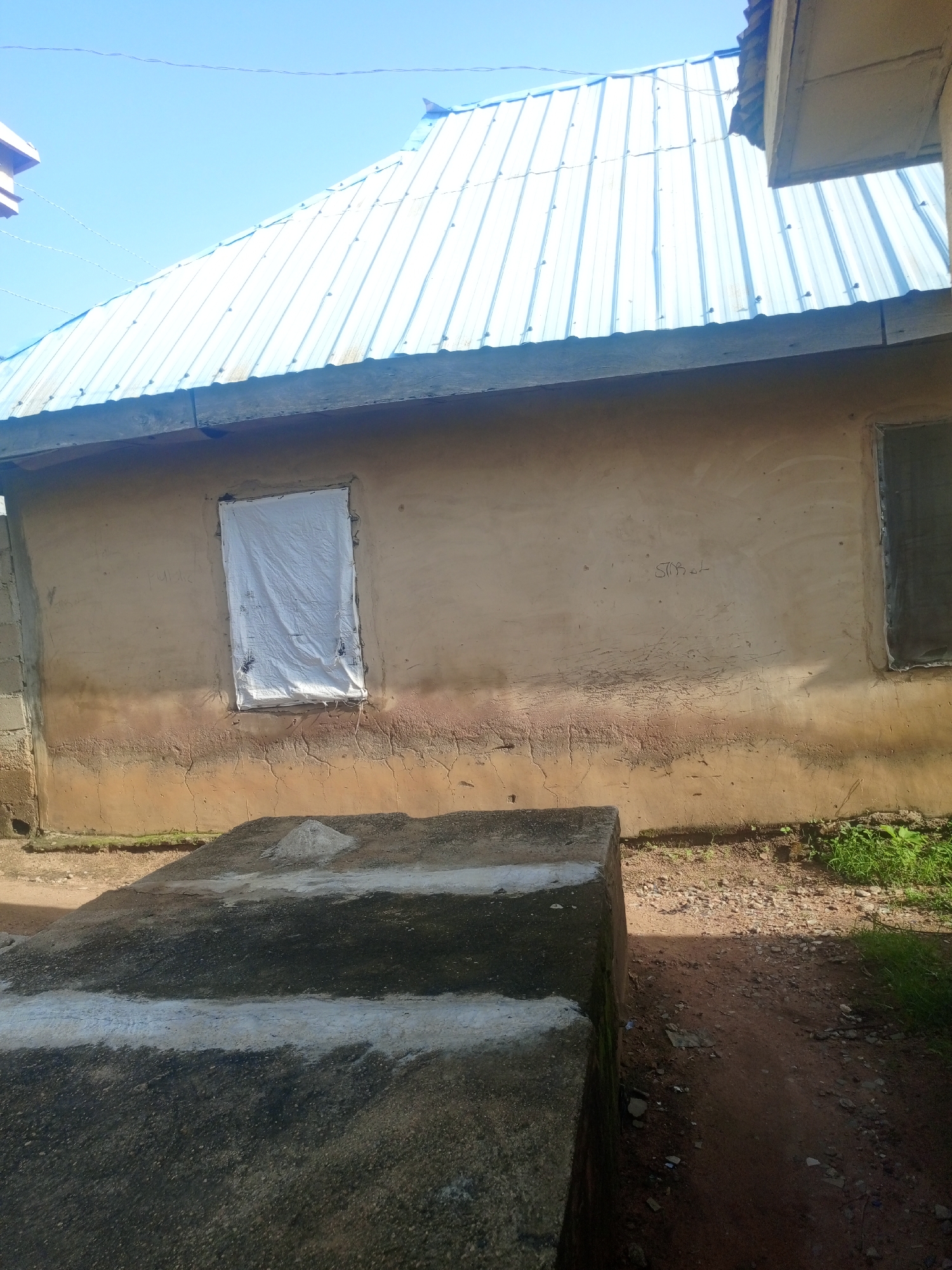 The image depicts the exterior of a building with a corrugated metal roof and beige-colored walls. One window is covered with a white tarp, and another appears to have a wire mesh. There is a concrete structure in the foreground, possibly a raised platform or steps, with some gravel and vegetation around it. The scene appears to be in an outdoor environment, with a clear blue sky above. The building's construction suggests it may be in a developing area. There are no people visible in the image. This scene is consistent with the typical architecture found in parts of Jalingo, Nigeria, particularly in residential or less developed areas.