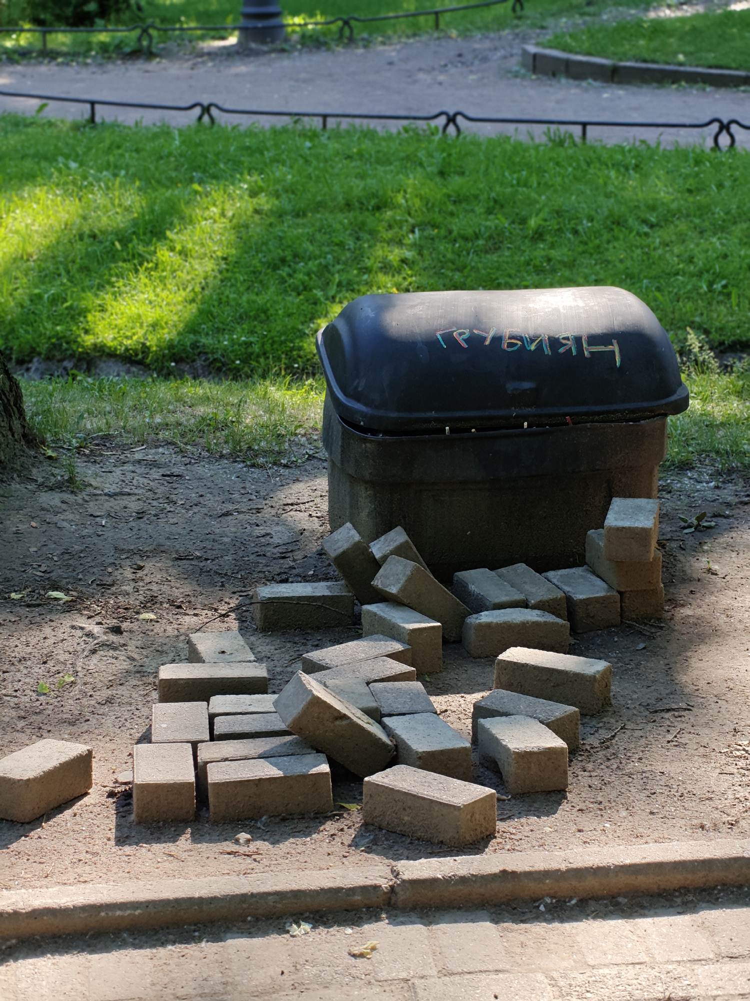 This media file captures an outdoor scene in Saint Petersburg, Russia. The primary subjects are a black dumpster with colorful graffiti on its lid, and a pile of concrete paving stones scattered on the ground in front of it. The dumpster appears to be made of dark, textured material and has a curved lid. The graffiti on the lid is in a Cyrillic script and is rendered in various bright colors, including green, blue, pink, and yellow.
The paving stones are rectangular and light gray, with some appearing dusty and worn. They are arranged in a somewhat disorganized manner, with some stacked and others lying flat. The ground is a mixture of dirt and sand, with patches of dry grass and some fallen leaves. To the left of the dumpster, a portion of a tree trunk is visible.
In the background, there is a patch of vibrant green grass, illuminated by bright sunlight, suggesting it is daytime and the weather is clear and sunny. Beyond the grass, a pathway is visible, bordered by a dark, decorative metal fence. A dark lamppost is also present in the distance, characteristic of park or street furniture in Saint Petersburg. A concrete curb runs along the bottom of the frame.
The scene suggests ongoing or recently completed landscaping or maintenance work, with the paving stones likely intended for repair or construction of pathways or other paved areas. The presence of the graffiti on the dumpster adds a touch of urban character to the otherwise naturalistic setting. The overall impression is a sunny day in a public space, possibly a park or courtyard, undergoing some form of renovation. The visible text on the dumpster reads "ГРУБИЯН", which translates to "RUDE PERSON" or "BULLY" in Russian.