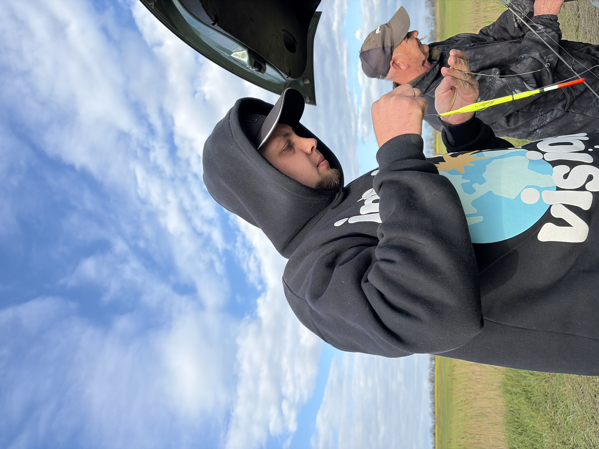The image shows two men fishing in Novomargaritovo, Russia. One man is wearing a black hooded sweatshirt with white lettering and a cap. The other man, older, is wearing a jacket and a baseball cap. He is holding a yellow and black fishing bobber. A vehicle with its trunk open is visible in the background. The sky is blue with scattered clouds. The foreground is a grassy field.