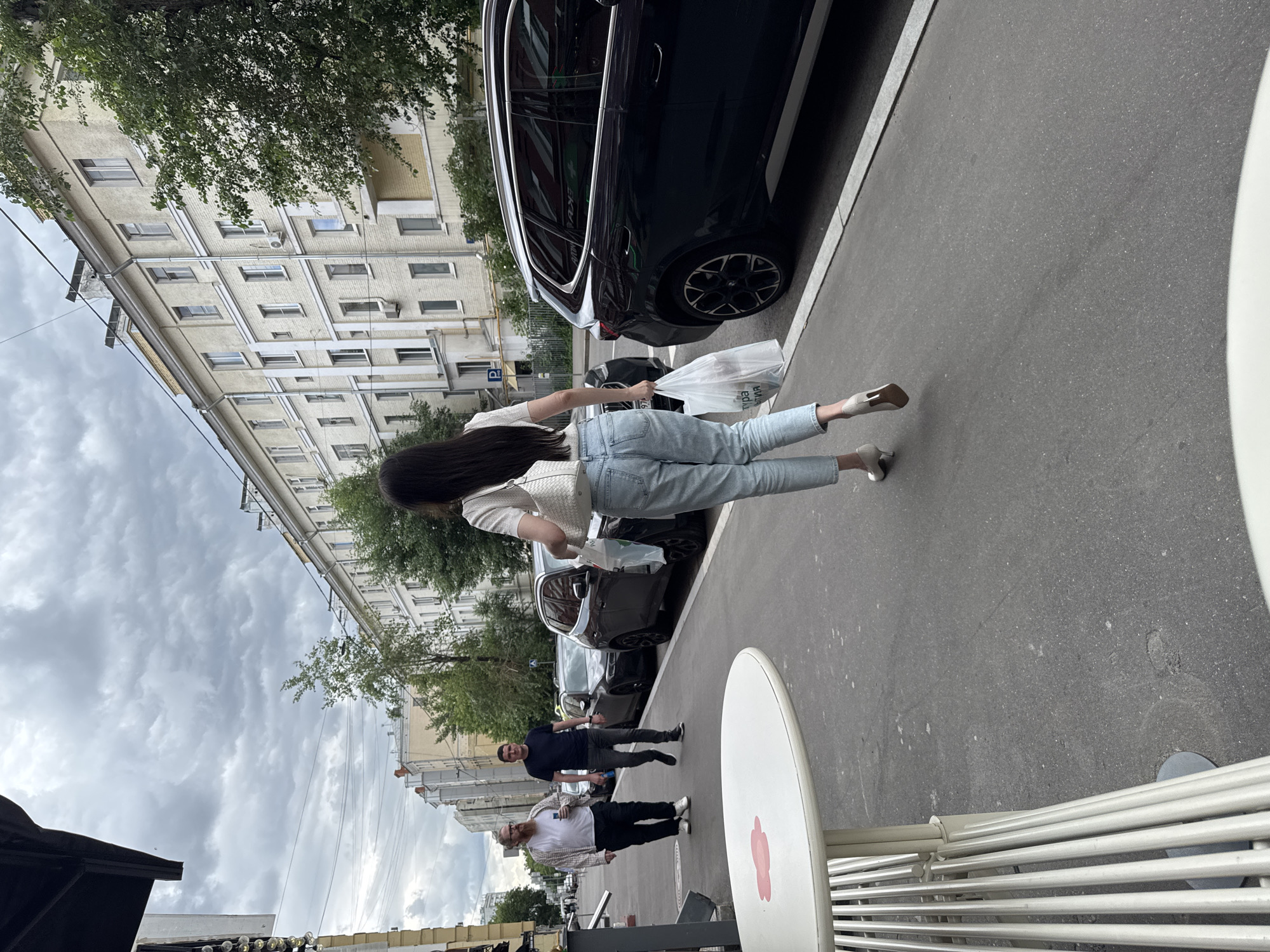 A woman with long, dark hair walks away from the camera down a city sidewalk in Moscow, Russia. She wears light blue jeans, a white short-sleeved shirt, and light-colored high heels. She carries a white woven purse on her left shoulder and a clear plastic bag in her right hand, which appears to contain white paper or plastic items. To her left, two men walk towards the camera. The man on the left has a red beard and wears a patterned shirt over a white t-shirt and dark pants. The man on the right wears a dark t-shirt and dark pants. They are walking on a gray asphalt sidewalk next to a street with parked cars. The buildings lining the street are multi-story, light-colored structures with numerous windows. The sky is overcast with gray clouds. A white outdoor table with a pink flower design is partially visible in the foreground on the left.