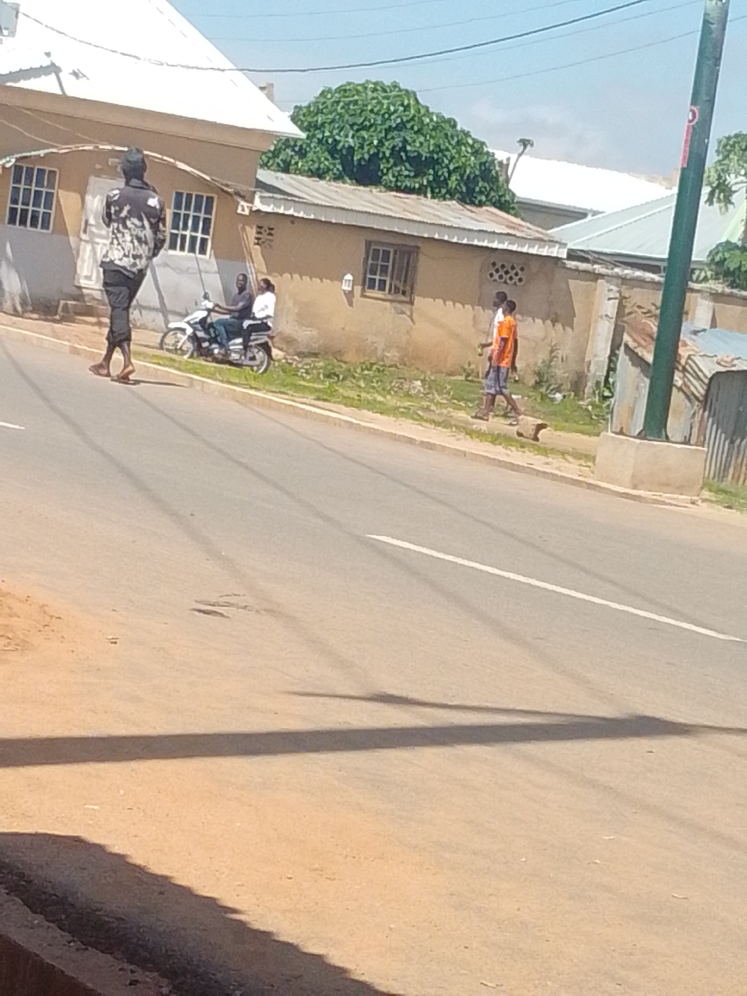In an outdoor urban scene, a person walks on a paved road in the foreground, casting a shadow. Behind them, a motorcycle with two occupants, a driver and a passenger, travels along the road. Further in the background, another person walks on the side of the road. Buildings with corrugated metal roofs and tan walls, along with some vegetation, are visible on the right side of the image, suggesting a potentially informal settlement or older urban area. A utility pole stands prominently on the right. The sky is clear and blue.