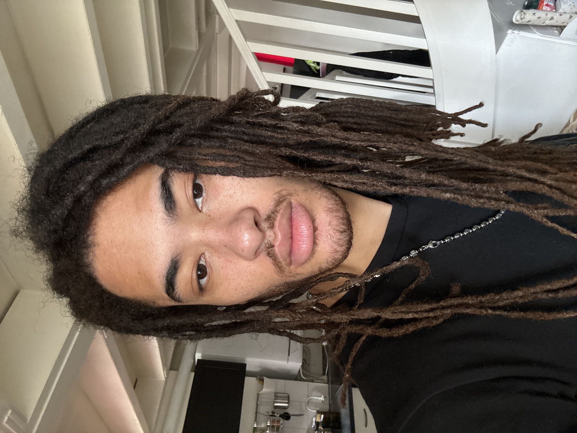 A close-up, slightly angled shot of a man with long dreadlocks. He is wearing a black t-shirt and a silver necklace. The background shows elements of an interior space including white banisters and a kitchen with stainless steel appliances. There is no evidence in the image to suggest a location in Paris, France.