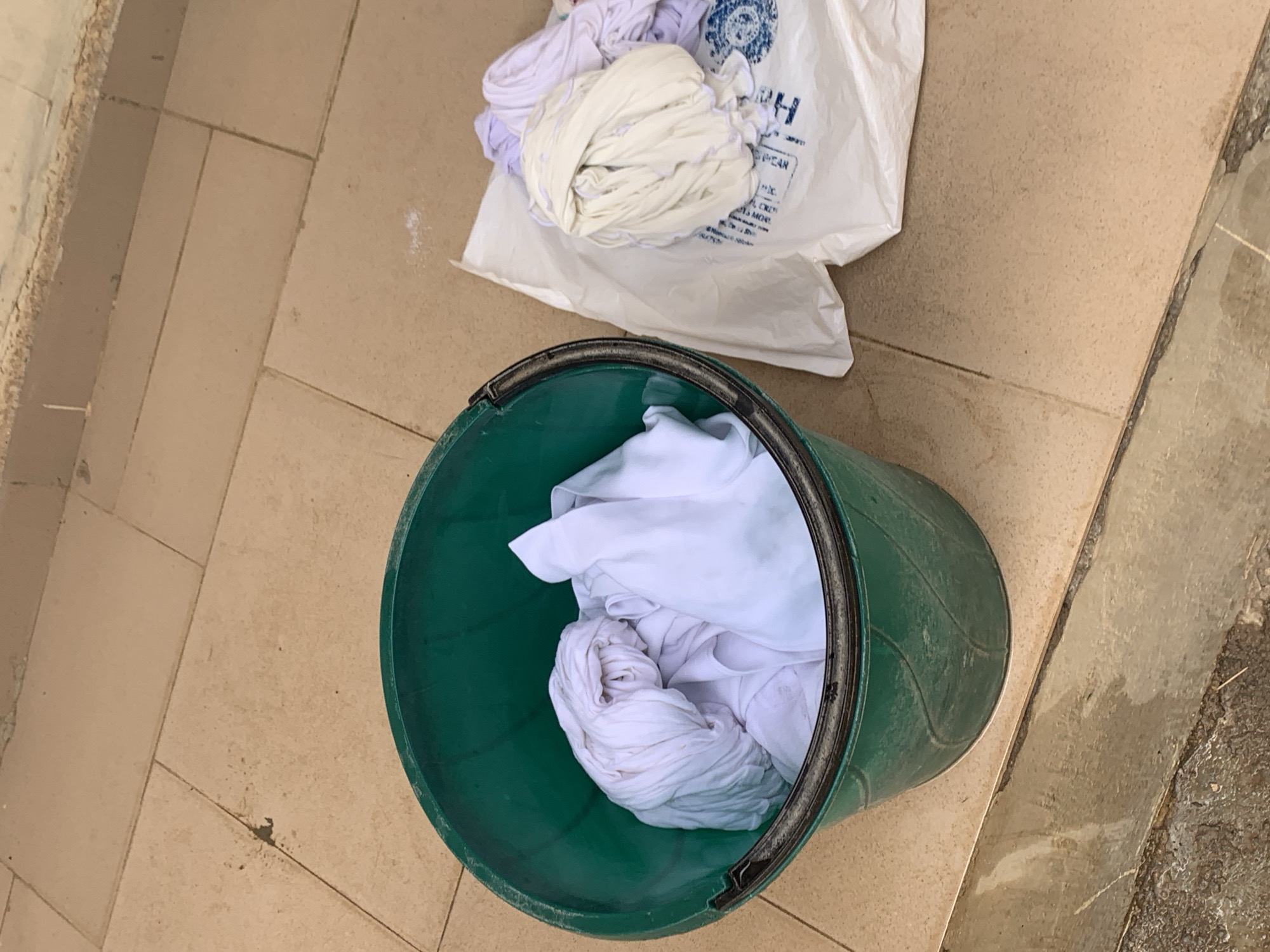 On a tiled floor, a green bucket is filled with crumpled white fabric. Next to the bucket, a white plastic bag filled with additional crumpled white fabric sits open. The location is presumed to be Ilorin, Nigeria. No people are visible. The primary objects are the bucket, bag, and fabric. The implied action is the collection of laundry. No interactions are directly observable. The environment appears utilitarian and domestic.