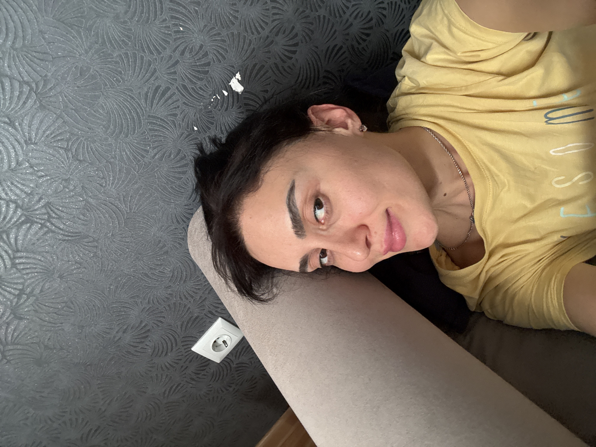 The image shows a woman with dark hair lying down, possibly on a couch or bed, with her head resting against the wall. She is wearing a yellow shirt and a silver necklace. The wall has a textured, swirling pattern and an electrical outlet is visible near her head. There is no indication of the city of Tbilisi, Georgia.