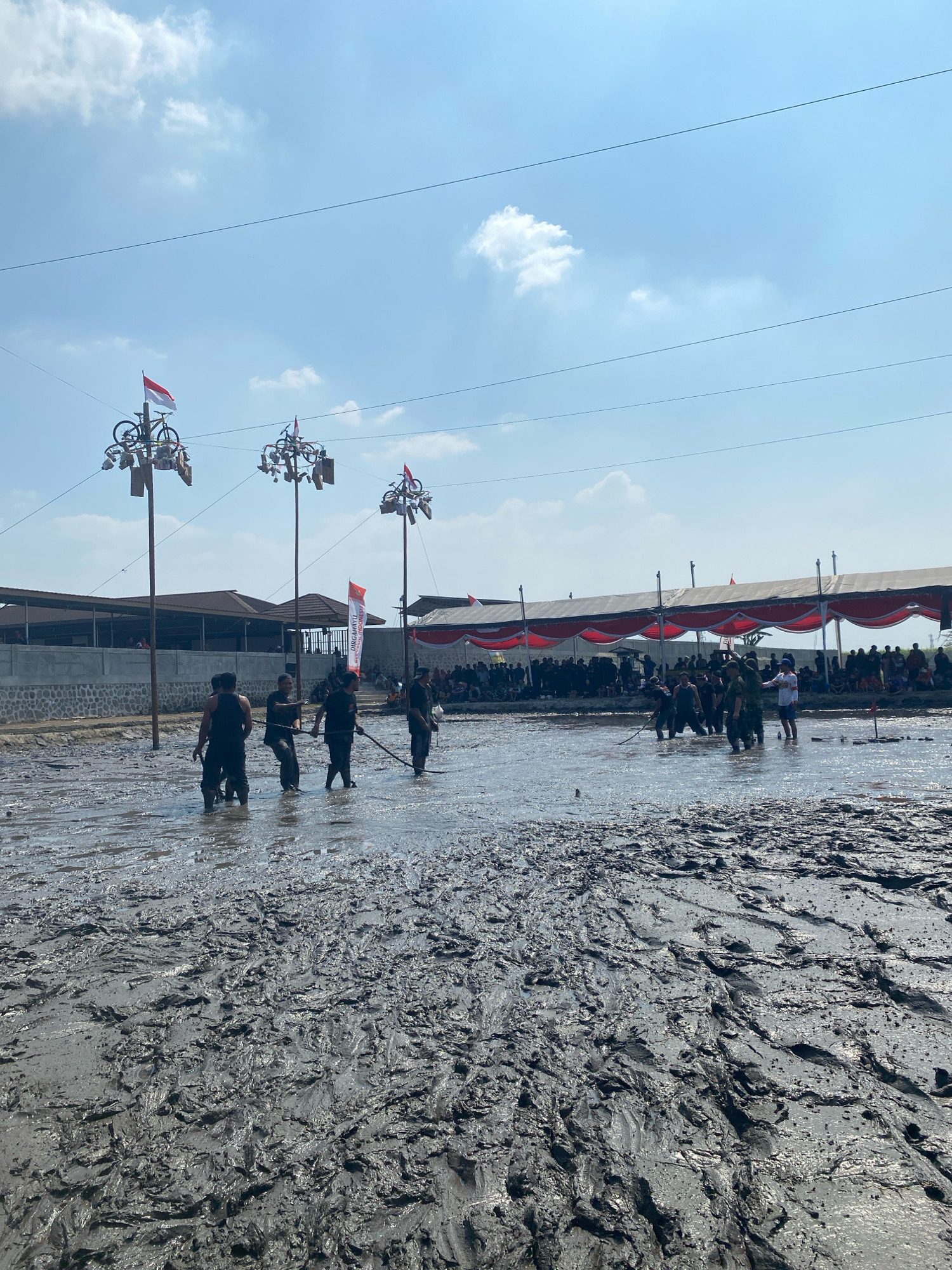 This image depicts an outdoor event in the city of Kediri, Indonesia, taking place in a muddy field under a bright, partly cloudy sky. In the foreground, the ground is covered in thick, wet mud with visible footprints and textured surfaces. Several individuals, dressed in casual dark clothing, are wading through the mud, some appearing to be participating in an activity or sport.
In the midground, tall poles are erected, each adorned with what appear to be bicycles and Indonesian flags. These poles are spaced out across the muddy area. Further back, a large gathering of spectators is visible, seated and standing behind a covered area with red and white draped fabric, suggesting a festive or ceremonial occasion. Buildings with pitched roofs and a stone wall are situated behind the poles and the crowd. Electrical wires stretch across the sky, indicating the presence of nearby infrastructure. The overall scene suggests a community event, possibly a traditional festival or competition, held in a rural or semi-rural setting within Kediri City.