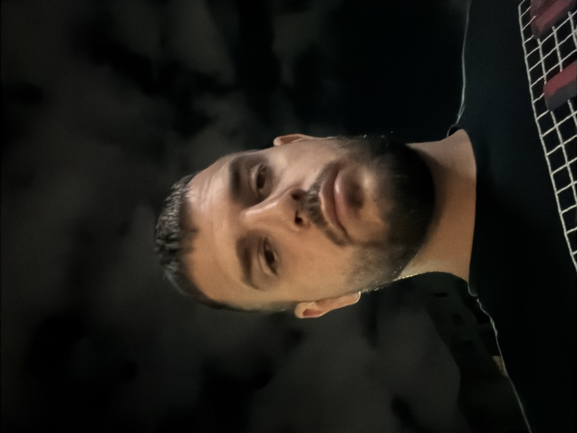 The image depicts a close-up, low-angle selfie of an adult male. He has short, dark hair, a trimmed beard, and fair skin, with a neutral expression, looking directly downward into the camera. He is wearing a dark, likely black, t-shirt with a white grid pattern on the chest, incorporating red rectangular or bar-like elements. The background is uniformly dark and indistinct, showing only subtle variations in shade, suggesting either a dark night sky with faint cloud cover or a very dimly lit interior with featureless dark walls. A direct, dim light source illuminates the subject from below or slightly in front. The location of the scene is in Sofia, Bulgaria.