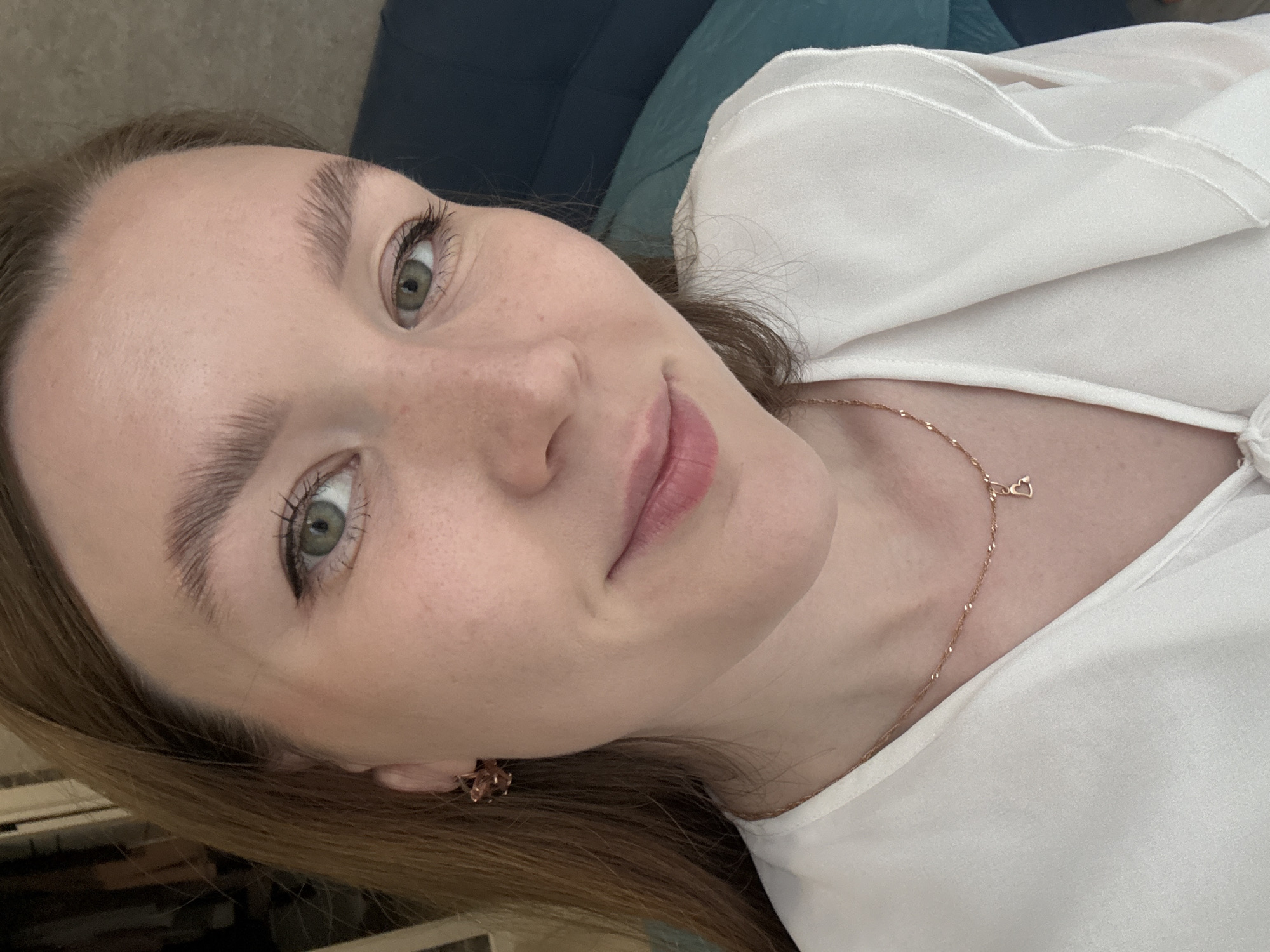 A close-up, slightly tilted portrait captures a young woman looking directly at the viewer with a soft, gentle expression and a hint of a smile. She has fair skin, light brown hair, and striking green eyes accentuated by long, dark eyelashes. Her eyebrows are full and brushed upwards. Some subtle freckles are visible across her nose and cheeks.
She is wearing a white blouse or top made of a soft, flowing fabric. A delicate gold-colored chain necklace with a small heart-shaped pendant adorns her neck. A small, intricate earring, possibly rose gold, can be seen in her right ear.
The background is softly blurred, revealing muted tones of dark blue and teal fabric, suggesting she is reclining on a couch or bed. The lighting appears soft and natural, indicating an indoor setting, likely during the day. No text is visible in the image. The scene conveys a sense of calm and peacefulness. The context provided indicates the location is Izhevsk, Russia.