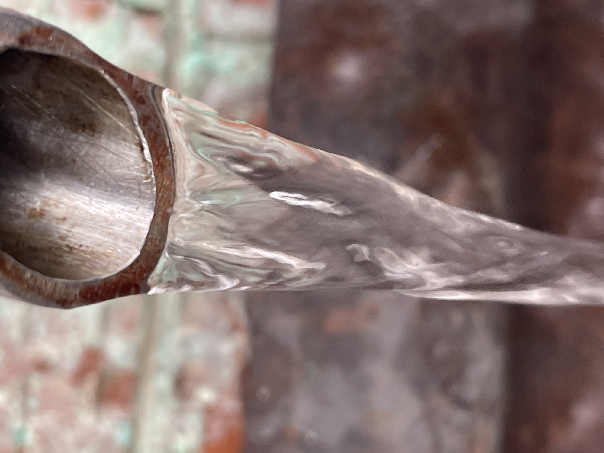 A close-up, slightly rotated, image shows water flowing from a pipe. The pipe is a rusty brown color, with a circular opening on the left side of the frame. The water is clear, with visible ripples and movement, filling the space between the pipe and the blurred background. The background consists of indistinct colors and shapes, with hints of green and brown, suggesting an outdoor setting. No people are visible.
