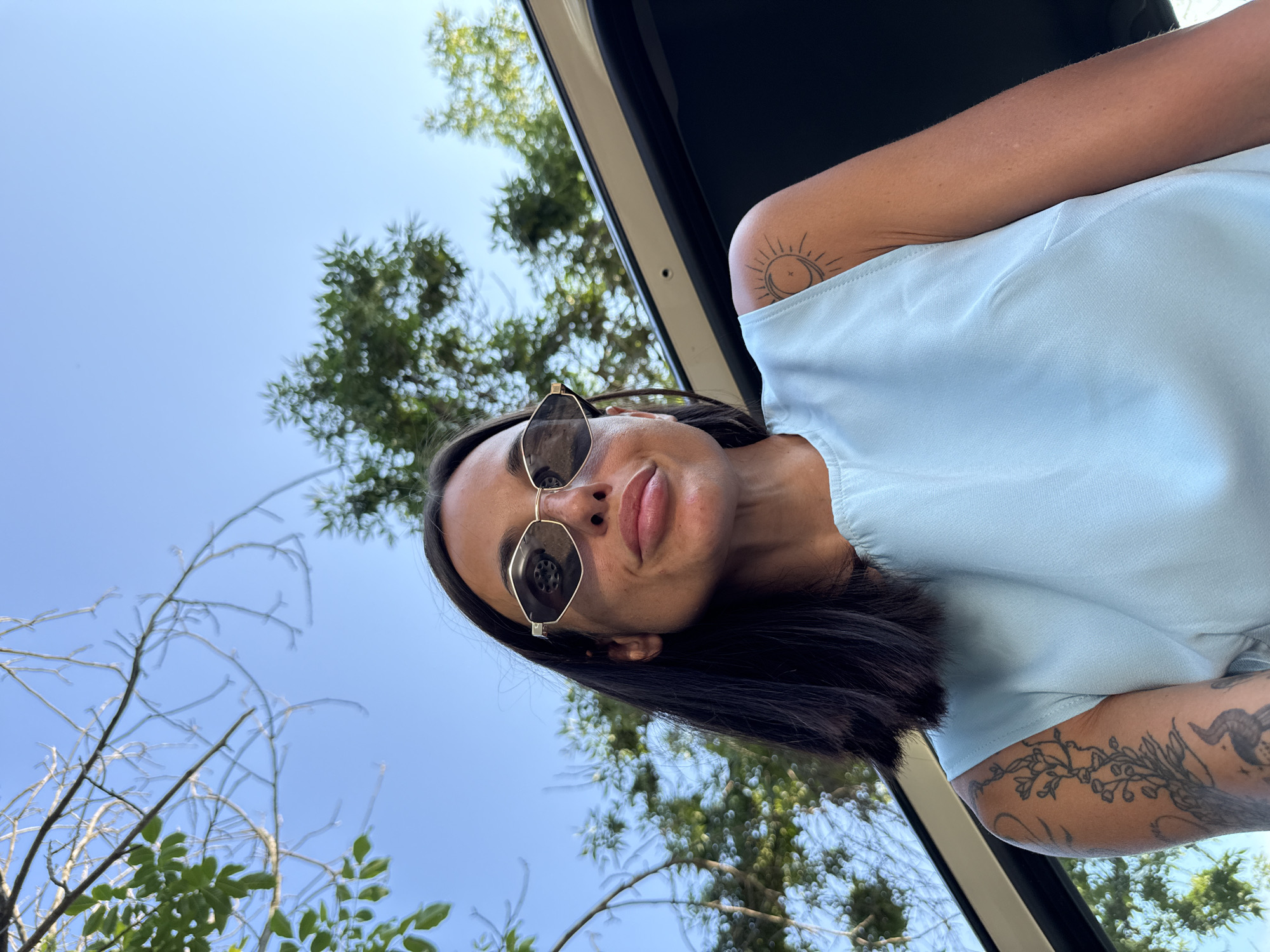 A low-angle, medium shot captures a woman looking slightly upward, positioned in front of a car door. She has dark hair, a fair complexion, and is wearing distinctive angular sunglasses with gold rims. Her lips are slightly parted, and she has a hint of a smile. She is wearing a light blue sleeveless top. Tattoos are visible on her left arm, including a floral design and a sun with a crescent moon on her right shoulder. The background consists of a clear blue sky and trees, suggesting an outdoor setting. The car door appears to be open, framing the woman. The image does not provide any specific visual cues to indicate a location in the city of Pleven, Bulgaria.
