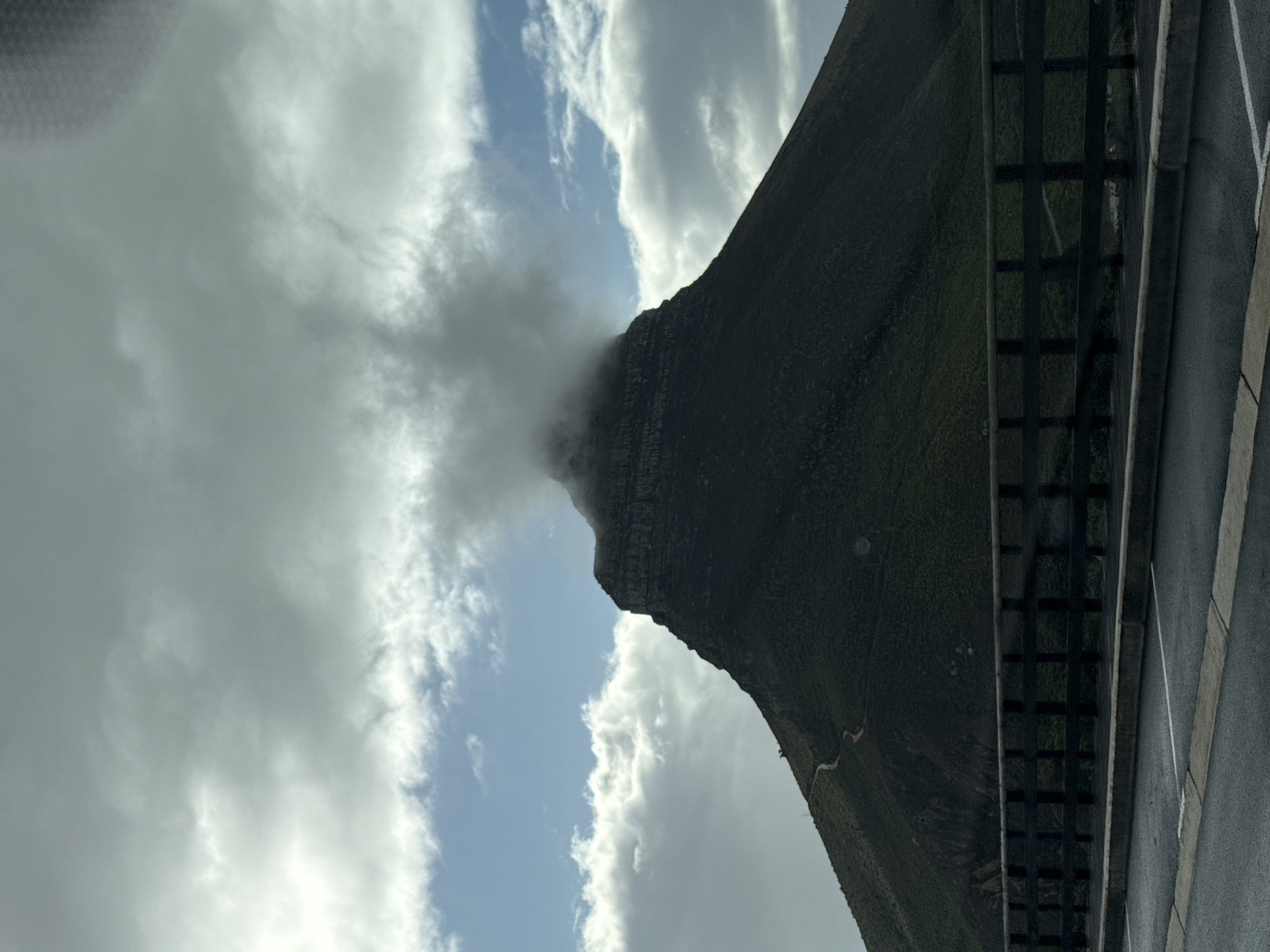 The image shows Lion's Head mountain in Cape Town, South Africa, its peak obscured by clouds. A roadway with a barrier runs along the bottom right of the frame. The sky is filled with a mix of white and gray clouds with patches of blue visible. The mountain is green and rocky, with a visible hiking trail winding up its slope. The photo is taken from an unusual angle, almost as if the photographer is tilted on their side.