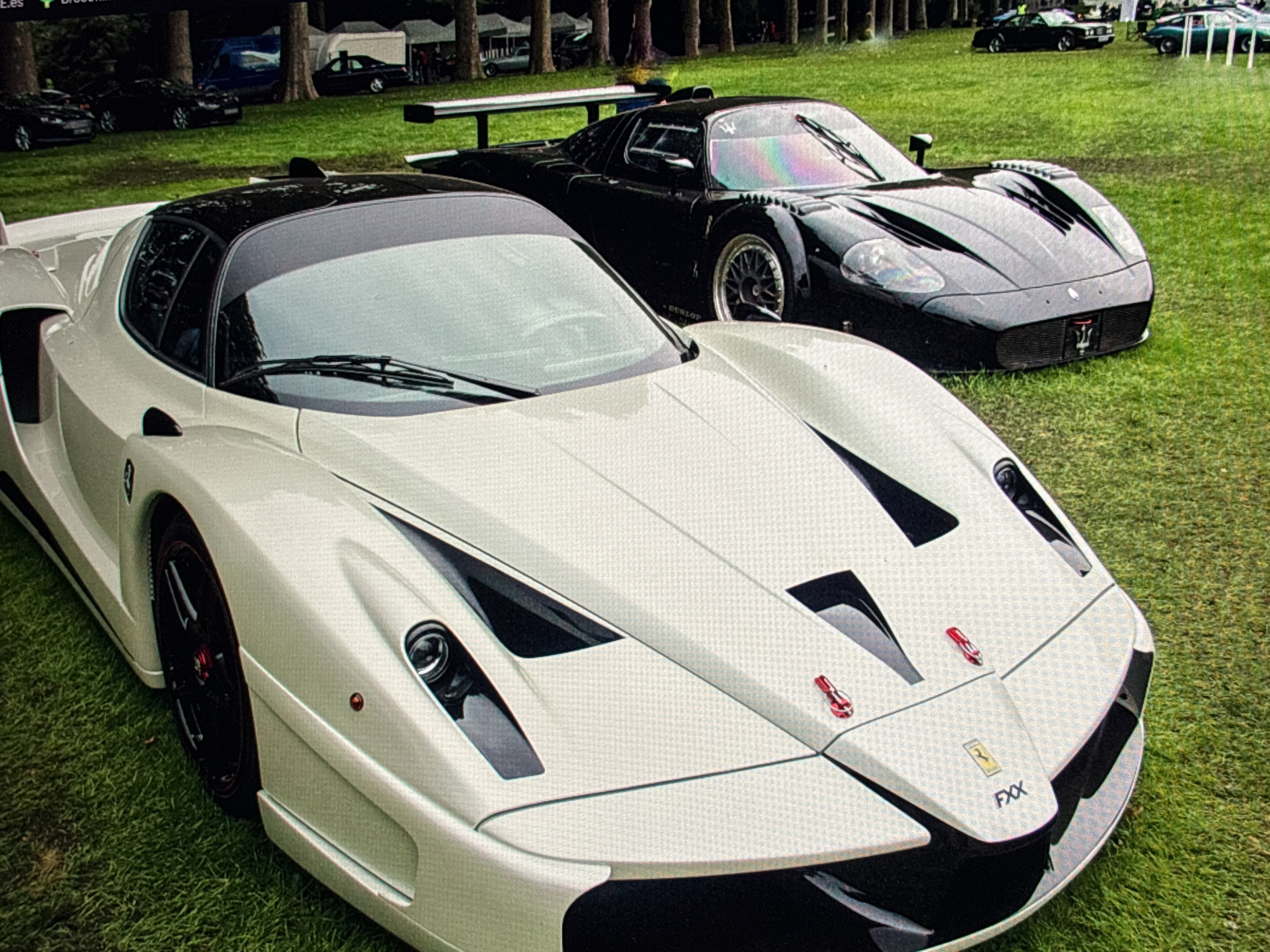 In an outdoor setting with a grassy lawn, two high-performance cars are parked. In the foreground, a white Ferrari FXX with a black roof and black wheels is partially visible, facing to the right. Its aerodynamic design, large spoiler, and prominent headlights are noticeable. Behind the white car, a black Maserati MC12 with a large rear wing and silver wheels is also parked. The Maserati's front is angled towards the right, revealing its aggressive styling. In the background, several other cars are parked on the grass, and trees line the perimeter of the area. Some tents and structures are also present in the distance. The overall scene suggests an automotive event or gathering. The image does not provide any specific details about the location in Sofia, Bulgaria.