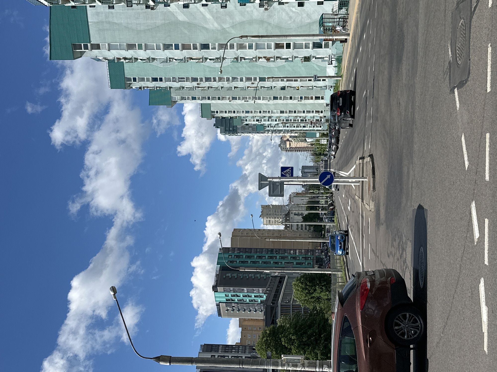 The image depicts a street scene in Minsk, Belarus, characterized by a wide road flanked by buildings under a partly cloudy sky. On the right side of the frame, a brown vehicle is parked near the curb. Further down the road, a blue car is visible amidst other parked cars. Road markings are present, delineating lanes, and street signs, including pedestrian crossing signs, are erected alongside the road. On one side of the road stand tall, light-colored buildings, likely apartment complexes, with numerous windows and a recurring green architectural accent. The opposite side of the street features a mix of modern buildings and possibly older structures further back. The sky above is a mix of blue and white, filled with cumulus clouds.