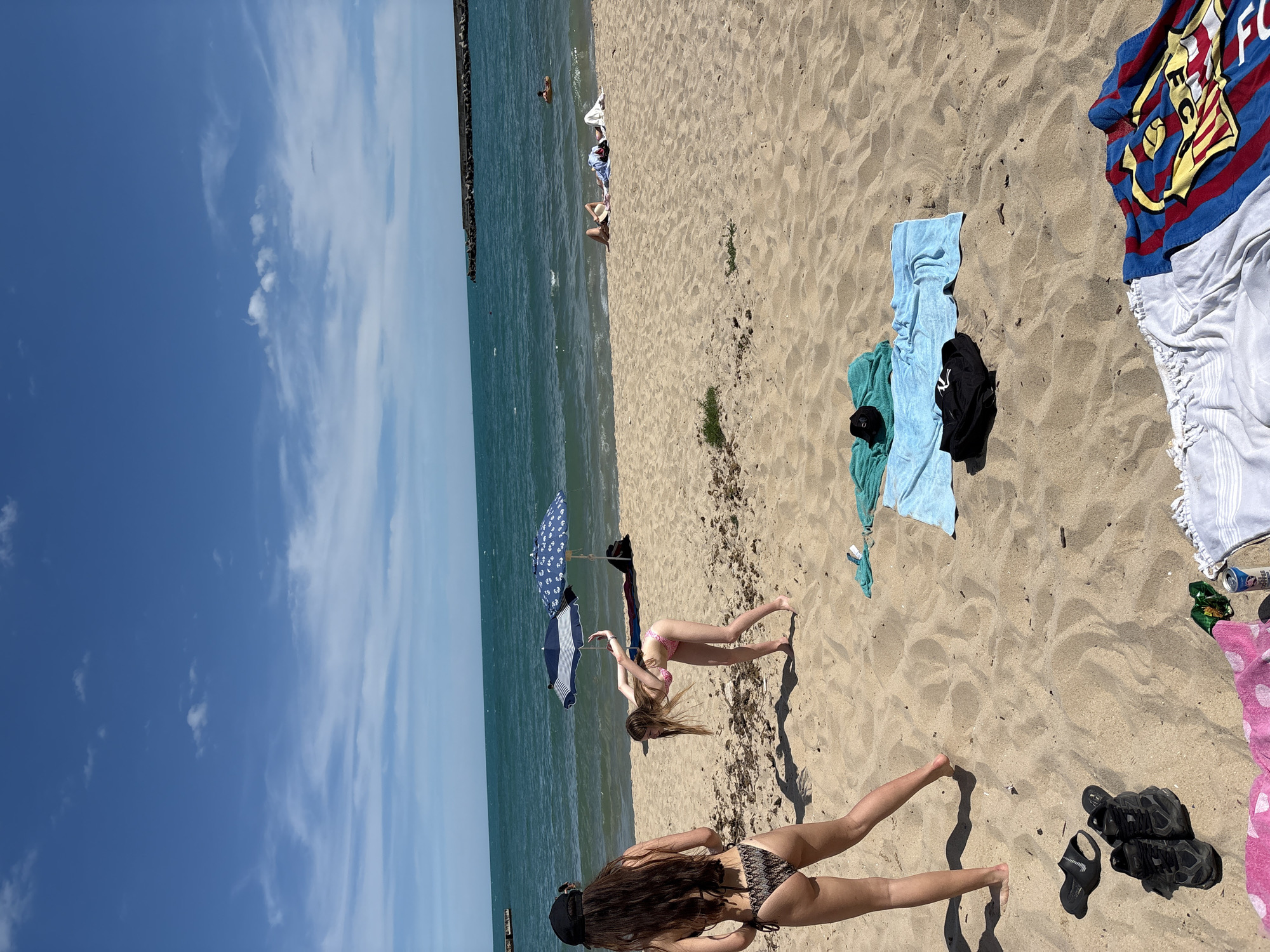 The image depicts a sunny day at a beach in Varna, Bulgaria. The scene is set on a sandy beach with the turquoise waters of the sea visible in the background, along with a breakwater. The sky is a vibrant blue with scattered white clouds.
The main subjects are two young women in bikinis. One woman is in the foreground, facing away from the camera, with her back to the viewer. She has long, dark hair and is wearing a patterned bikini. The second woman is further back on the beach, also facing away, in a pink bikini. She is in a yoga pose, with her arms outstretched. A closed umbrella with a blue and white pattern stands beside her.
Several objects are scattered on the sand, including towels of various colors (blue, light blue, white, and a brightly colored towel with red, blue, and yellow stripes), a black jacket, a black baseball cap, and a pair of black sandals. There is also a can of what appears to be a beverage.
The overall atmosphere is relaxed and leisurely, typical of a beach day. The lighting suggests it is daytime, likely mid-morning or afternoon, given the shadows cast on the sand. There is no visible text in the image.