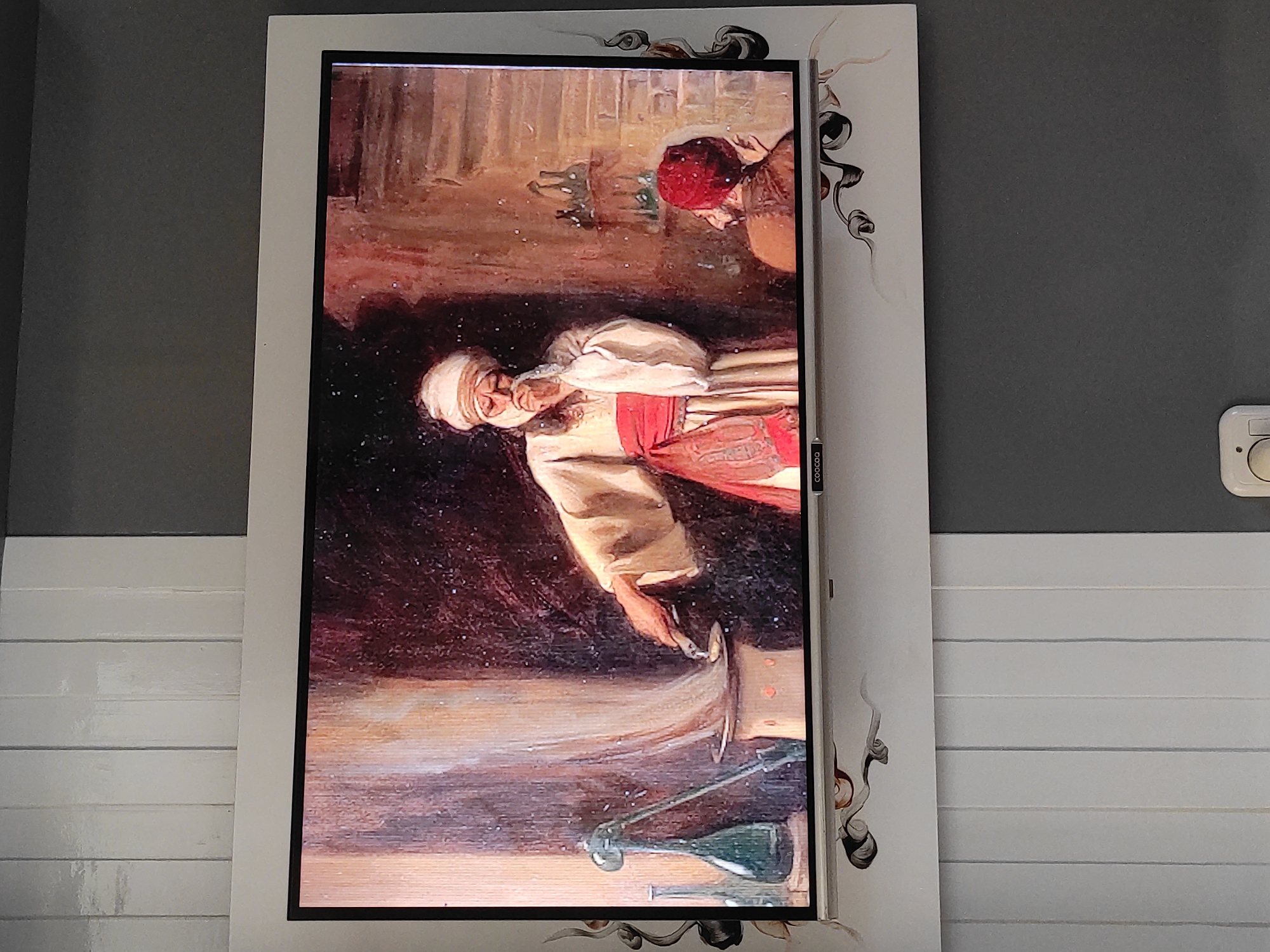 The image shows a television screen displaying a painting of two men in what appears to be an alchemist's laboratory. The foreground on the screen features a man in traditional Middle Eastern attire, including a turban and a red sash, bending over a glowing apparatus from which smoke is rising. He is holding a tool and seemingly manipulating a substance. To his right, another man wearing a red fez sits observing. The background shows shelves filled with bottles and jars, suggesting a setting for scientific experimentation or potion-making. The television is mounted on a wall decorated with vertical white panels and a dark grey section. There are decorative, abstract swirl patterns on the white frame of the television. The location in Sumedang, Indonesia, is not evident from the image itself.