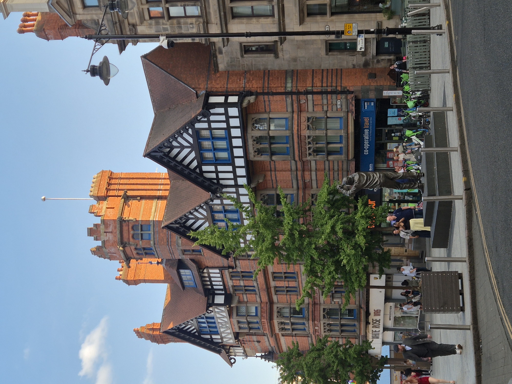 The image shows a street scene in Nottingham, United Kingdom, featuring a statue in the foreground and a distinctive brick building with half-timbered elements in the background. The building is an example of Victorian architecture. A statue of a man with his arms raised is positioned on a raised platform. In front of the building, there are several people walking, some standing, and a row of electric scooters. A sign for "CO-OPERATIVE TRAVEL" is visible on the building. The street is paved, and there are lampposts and bollards visible. The sky is blue with some clouds. The overall impression is of a bustling urban environment on a clear day.