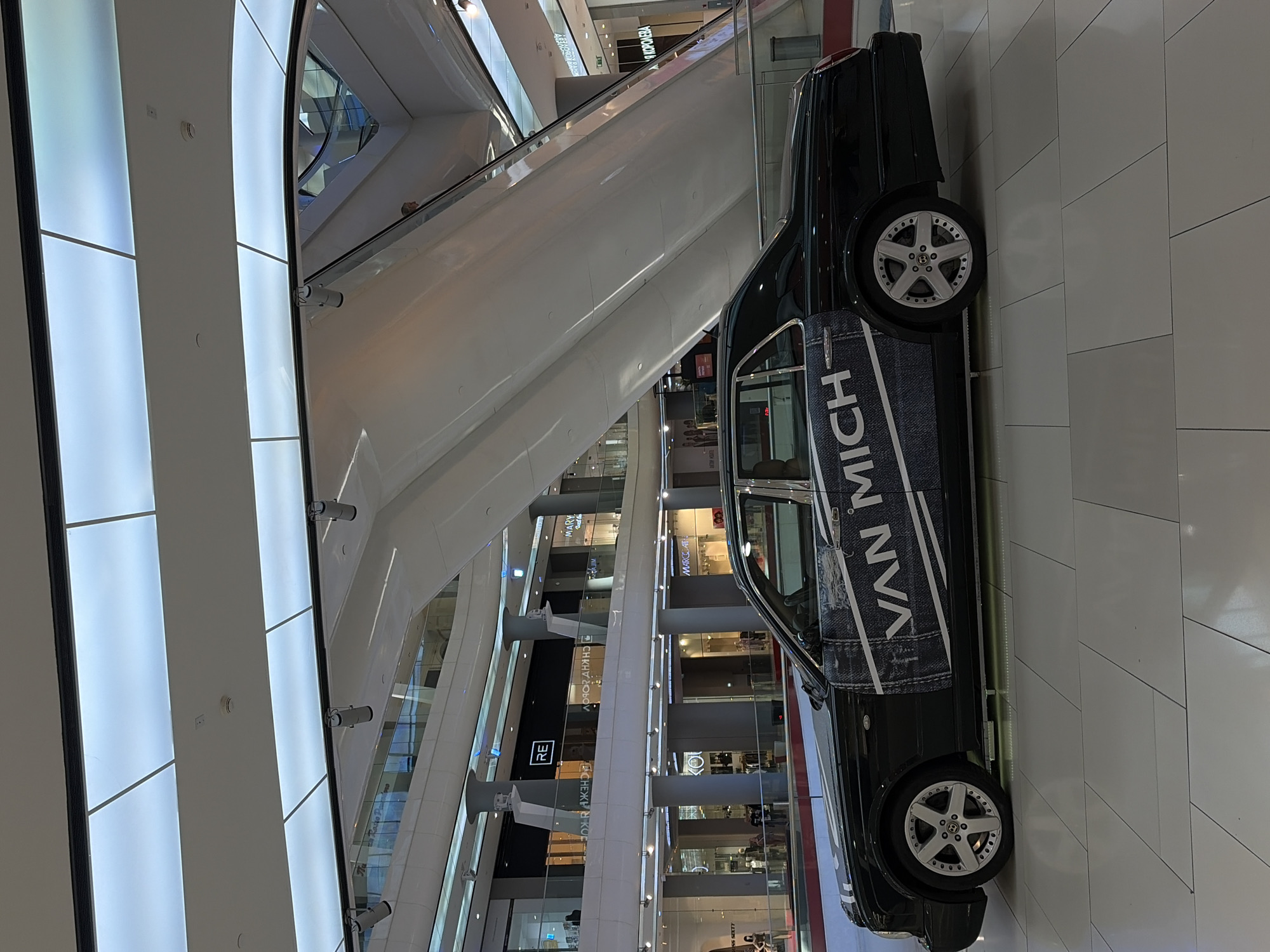 An indoor shot captures a multi-story shopping mall in Krasnodar, Russia, showcasing modern architecture and retail spaces. The image is taken from a slightly elevated perspective, looking down at the ground floor and across several levels.
The main subject is a dark-colored sedan, possibly black or dark green, prominently displayed on the ground floor. Its side door features a distinctive decal with "VAN MICH" written in large white letters against a blue denim-like texture. The car is equipped with multi-spoke alloy wheels.
In the background, escalators ascend through the bright, airy space. One person is visible on an escalator, moving upwards. The mall's interior features sleek white walls, glass railings, and polished light-colored floor tiles. Large diffused light panels illuminate the upper walls and ceiling, creating a very bright environment.
Numerous retail store fronts are visible on various levels, featuring large glass windows. Visible text includes "VAN MICH" on the car, "RE" on a store facade, and several Cyrillic signs such as "СНЕЖНАЯ КОРОЛЕВА" (Snezhnaya Koroleva - Snow Queen), which is a well-known Russian fashion chain. Other partial or reversed text snippets include "CHKHOLOV," "MARY," and "MAROCCAT" (likely Marocain or similar). A fire alarm or smoke detector is mounted on a white wall panel. The overall impression is that of a clean, contemporary commercial space.