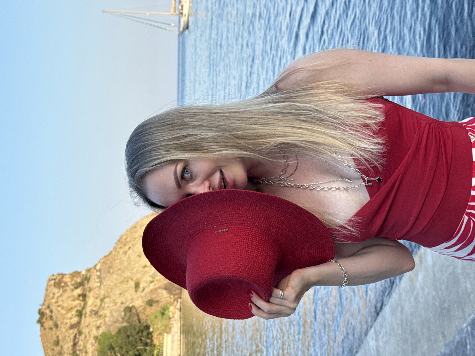 A woman with blonde hair is standing on a stone embankment by the sea, holding a large red hat in front of her face, obscuring most of it. She is wearing a red top and a skirt with red and white stripes. She has on a silver bracelet and necklaces. In the background, there is a rocky cliff face, trees, and a sailboat on the water. The sky is clear and blue. The location is the city of Utios, Ukraine.