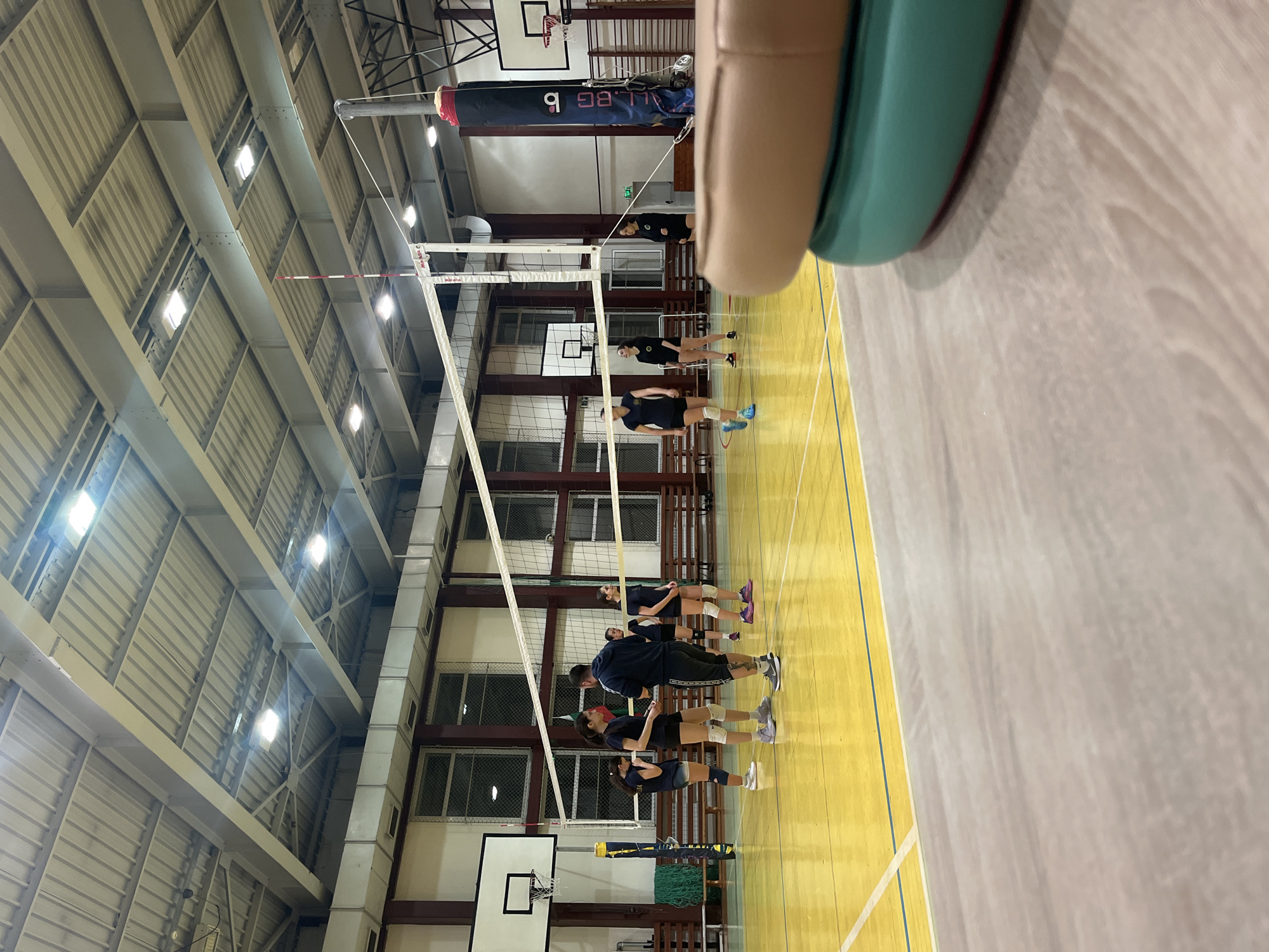 A gymnasium in Sofia, Bulgaria, is set up for volleyball. A group of people are on the court, likely practicing or warming up. The court is yellow, with white and green lines marking the boundaries. A volleyball net stretches across the center of the court. A basketball hoop is visible in the background. The gym has a high ceiling with metal supports and several windows. A person's arm, perhaps the photographer, is in the foreground.