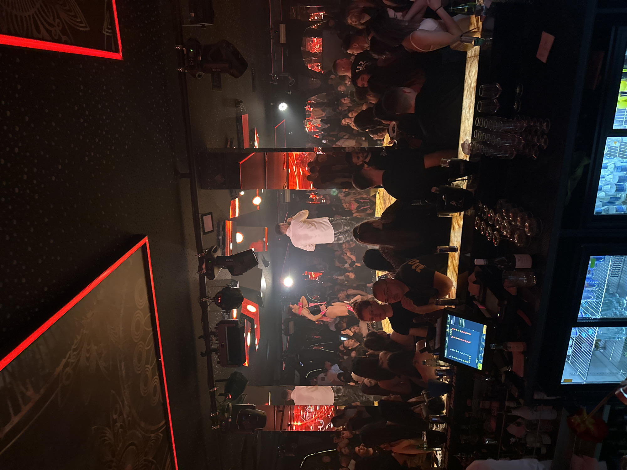 The image depicts a crowded nightclub in Sofia, Bulgaria. People are dancing and mingling in a dimly lit space with red lighting accents. A performer in a pink outfit lies on a raised platform, seemingly the focus of attention. A DJ booth is visible with a control panel and computer screen. Behind the booth, a bar with glasses and bottles is present, adjacent to a refrigerator stocked with bottled beverages. The overall environment suggests a lively nightlife setting.