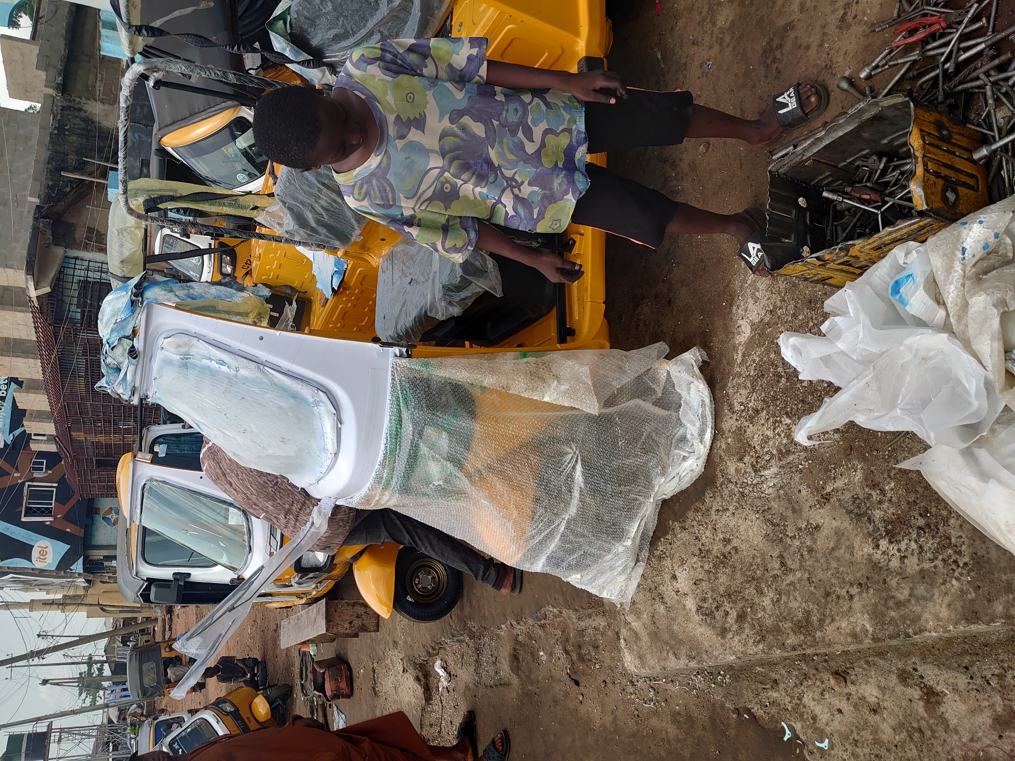 The image shows several new, yellow three-wheeled vehicles parked outside a building. One vehicle has a white cab door partially open. A young person in a patterned shirt and dark shorts is standing near one of the vehicles. Next to the person is an open toolbox filled with tools. The ground is concrete with some scattered debris and plastic bags. The location appears to be in an urban area.