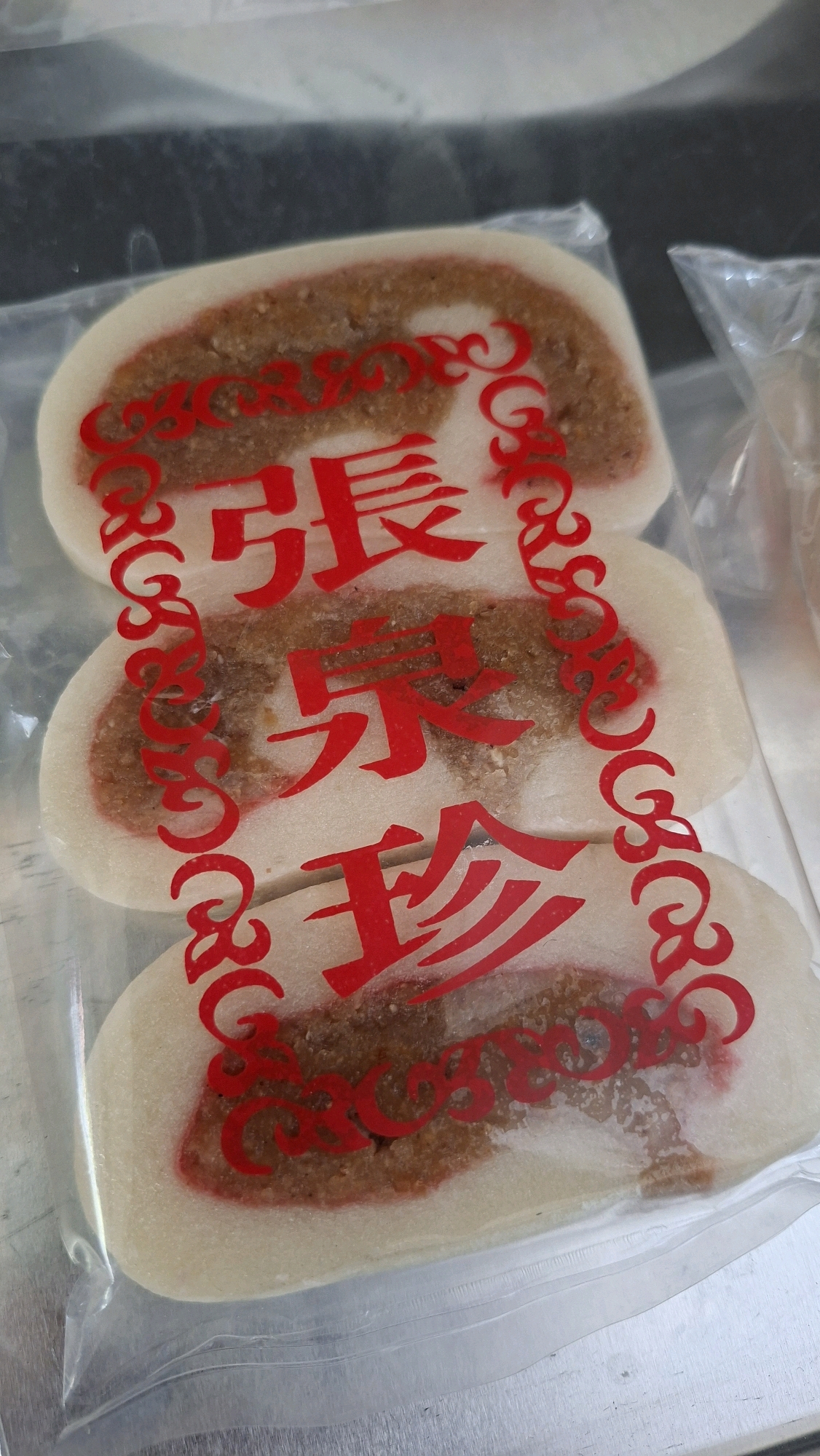 The image shows three pieces of sweet bread, stacked and wrapped in clear plastic. Each piece is oval-shaped and has a white outer layer with a reddish-pink border. The center of each piece is filled with a brown, granular substance, possibly nuts or seeds. Red Chinese characters are printed vertically on the plastic wrap, superimposed over the bread. The characters appear to be "張", "泉", and "珍". The bread is on a metallic surface, possibly a display case or counter. The overall impression is that of a food item for sale. No people are visible in the image. The location is not specified in the image.