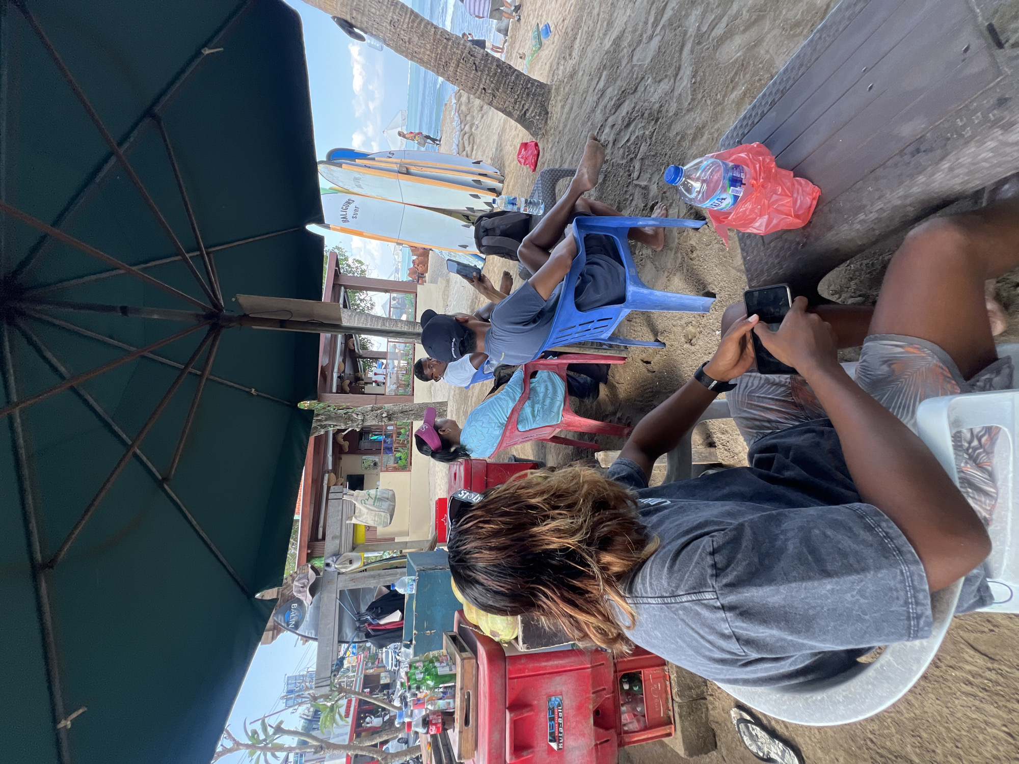 The image shows a group of people under a large green umbrella on a sandy beach in Kuta, Indonesia. In the foreground, a person with long, wavy brown hair sits in a white chair, holding a mobile phone and wearing a gray t-shirt and patterned shorts. To their right, a blue plastic chair is occupied by a person in a dark t-shirt and shorts, also looking at a mobile phone, with a bottle of water in a red plastic bag on a wooden table. Behind them, a woman with a pink visor sits in a red chair, wearing a light blue top. Further back, other people are visible, and surfboards are propped up against a palm tree. The background shows the ocean, the sky with clouds, and some buildings with shops and stalls, typical of the Kuta beachfront. The scene appears relaxed, with people taking a break from beach activities.