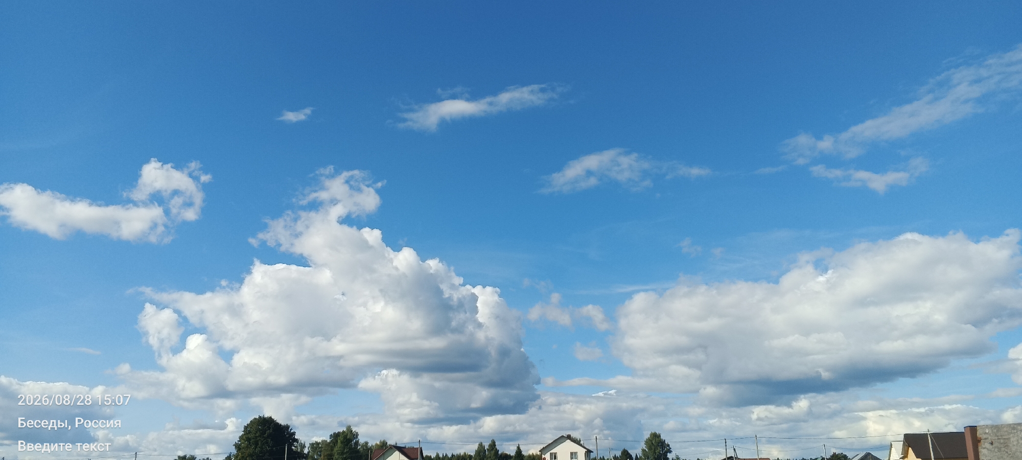 The image captures a daytime view of a sky filled with white and gray cumulus clouds against a clear blue background. In the lower portion of the frame, the tops of trees, houses, and utility poles are visible, indicating a rural or suburban setting. A timestamp and location, "2026/08/28 15:07" and "Besedy, Russia" are overlaid on the bottom left corner of the image, along with the text "Введите текст" (Enter text). The overall scene suggests a serene, natural environment with signs of human habitation.