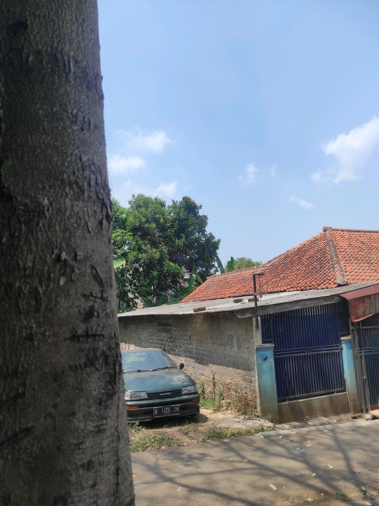 A dark-colored Daihatsu car with license plate D 1428 SM, dated 07-27, is parked on a dirt and grassy area next to a grey brick wall. A large tree trunk is in the foreground on the left. Behind the car, a blue metal gate with a decorative iron crossbar is visible, with light blue support pillars. Beyond the gate is a building with a terracotta tile roof. Green trees and a bright blue sky with scattered white clouds complete the scene, suggesting a daytime outdoor setting in Padalarang, Indonesia.