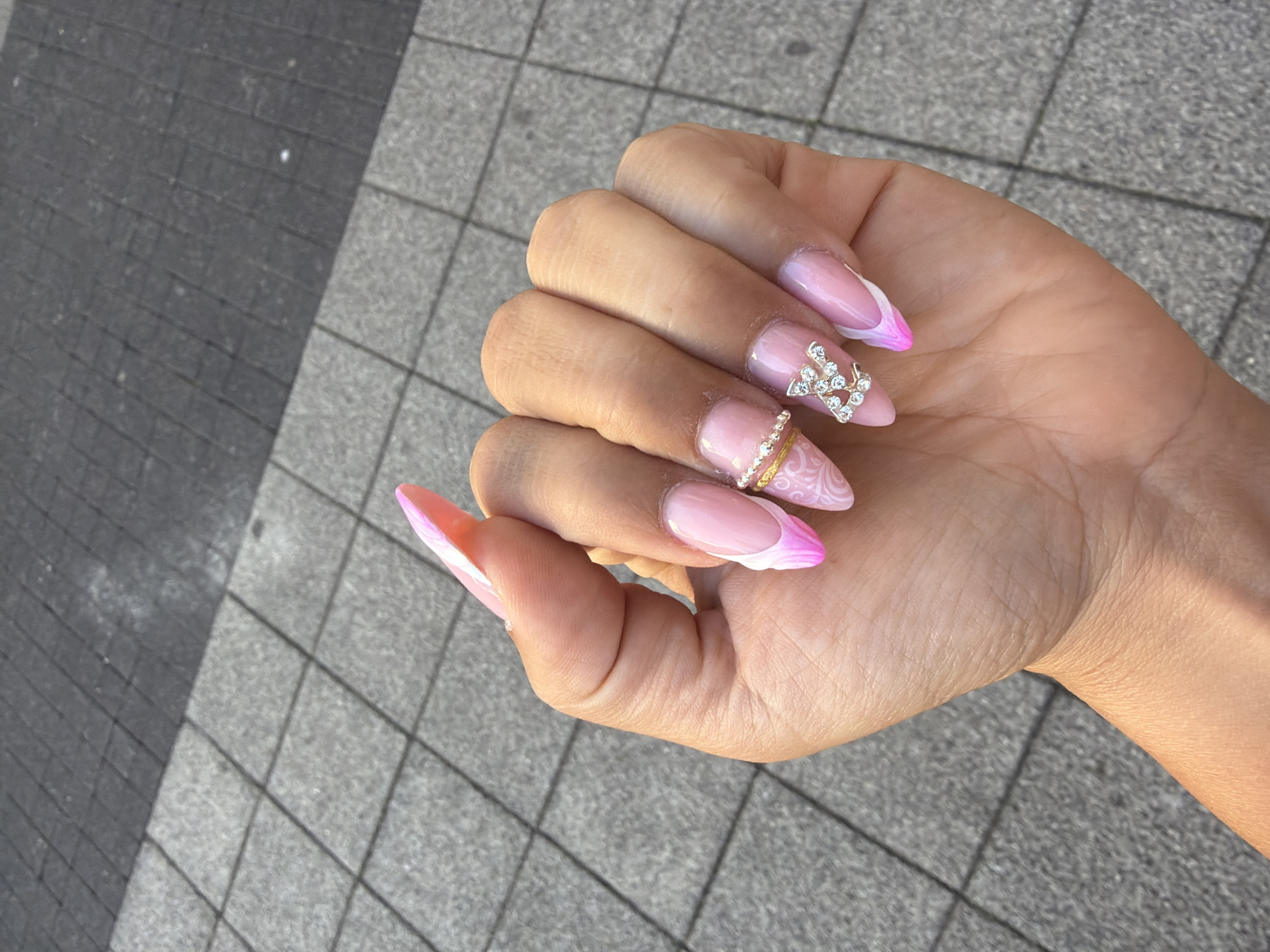 A close-up, overhead shot shows a person's left hand with manicured nails resting on a grey, tiled outdoor surface in Schwelm, Germany. The nails are long, stiletto-shaped, and feature a light pink French manicure with white and darker pink detailing. The index finger nail has a delicate swirling pattern and is adorned with a thin, gold-colored band made of small rhinestones. The middle finger nail is decorated with a silver-colored "LV" monogram made of clear rhinestones. The person's skin appears tanned. The background is a blurred view of the grey tiled ground, suggesting an urban outdoor environment.