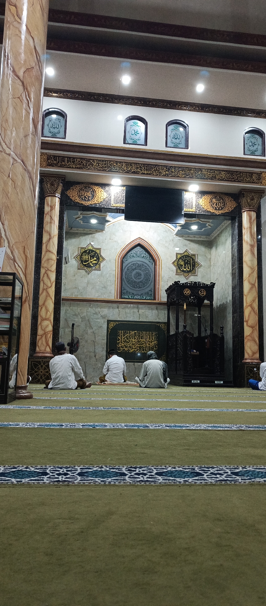 In the interior of a mosque in Arjasa, Indonesia, three individuals are seated on the floor, facing a decorative wall featuring Islamic calligraphy. The individuals are all males, dressed in white, seated in a prayer posture with their backs to the camera. The mosque interior is adorned with ornate Islamic calligraphy in various frames and designs on the walls. A black wooden pulpit stands to the right of the central wall. The floor is covered with a green carpet with a patterned border. The overall lighting suggests an indoor setting.