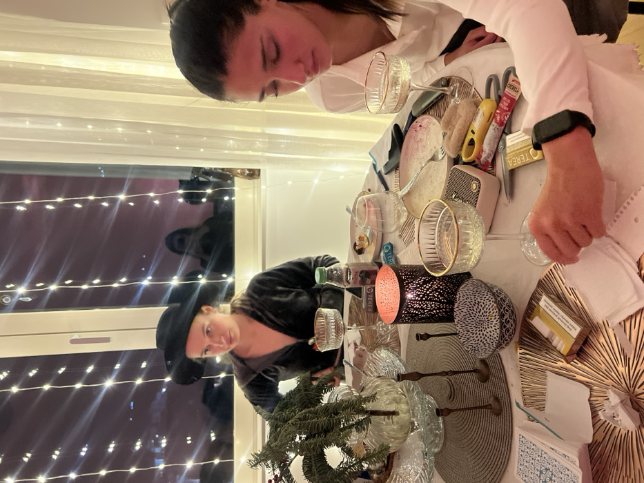Two women are seated at a table. One wears a white shirt, and the other has on a black velvet jacket and hat. The table is laden with glasses, plates, and a small Christmas tree. A speaker, cigarette packets, and cards are also visible. Lights hang in the background, visible through a window in which a reflection is seen.